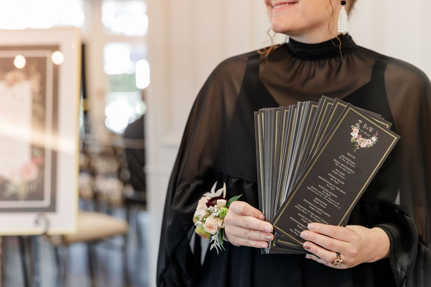 Woman in a black sheer blouse with a floral wrist corsage holds a stack of dark printed wedding menus indoors.