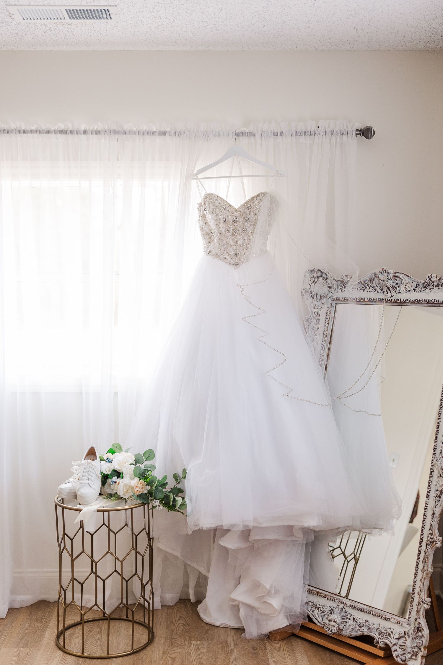 White wedding dress with beaded bodice hanging near an ornate mirror and floral arrangement.