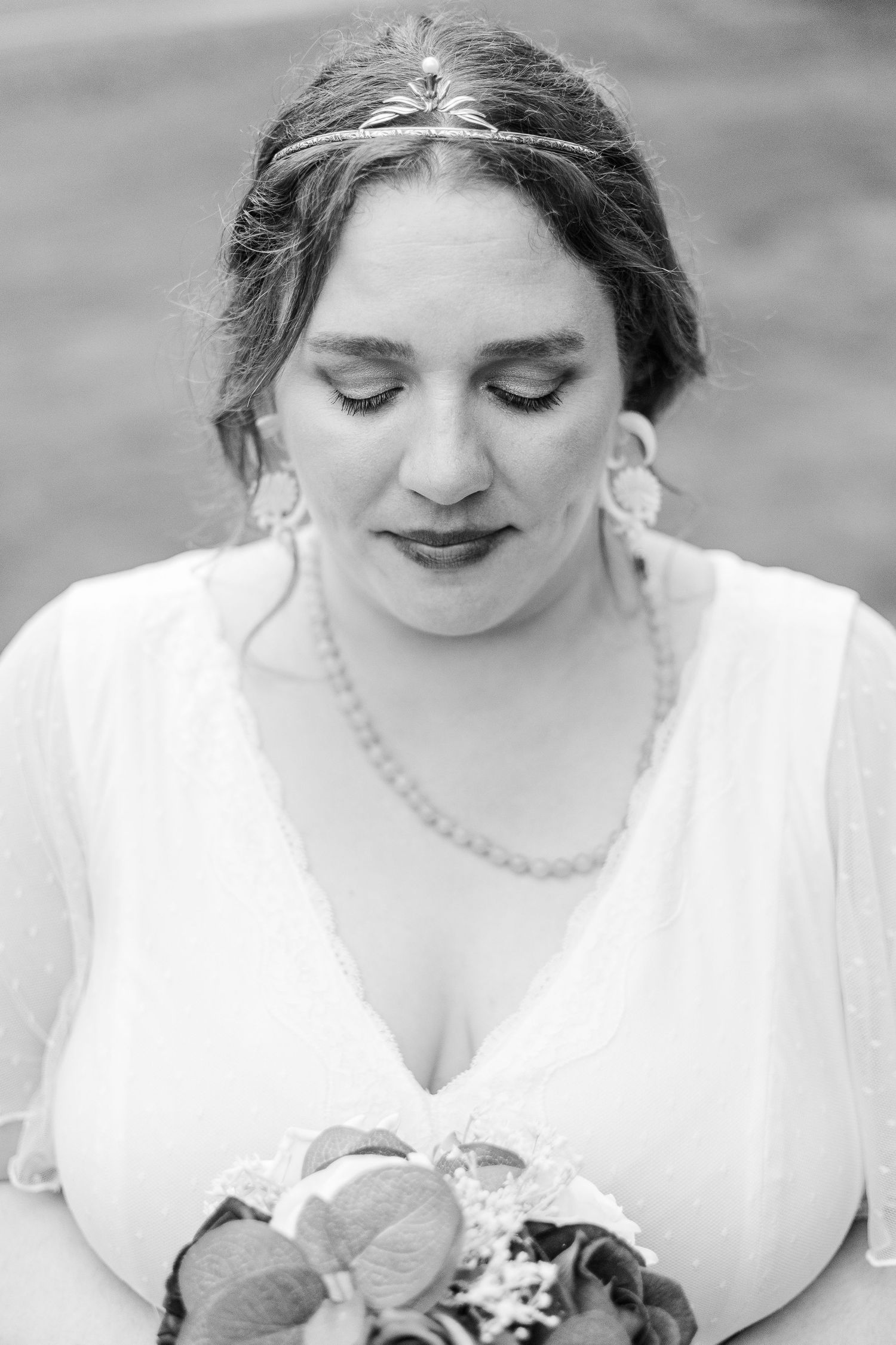 Bride in white dress with delicate jewelry gazes down in classic black and white portrait.