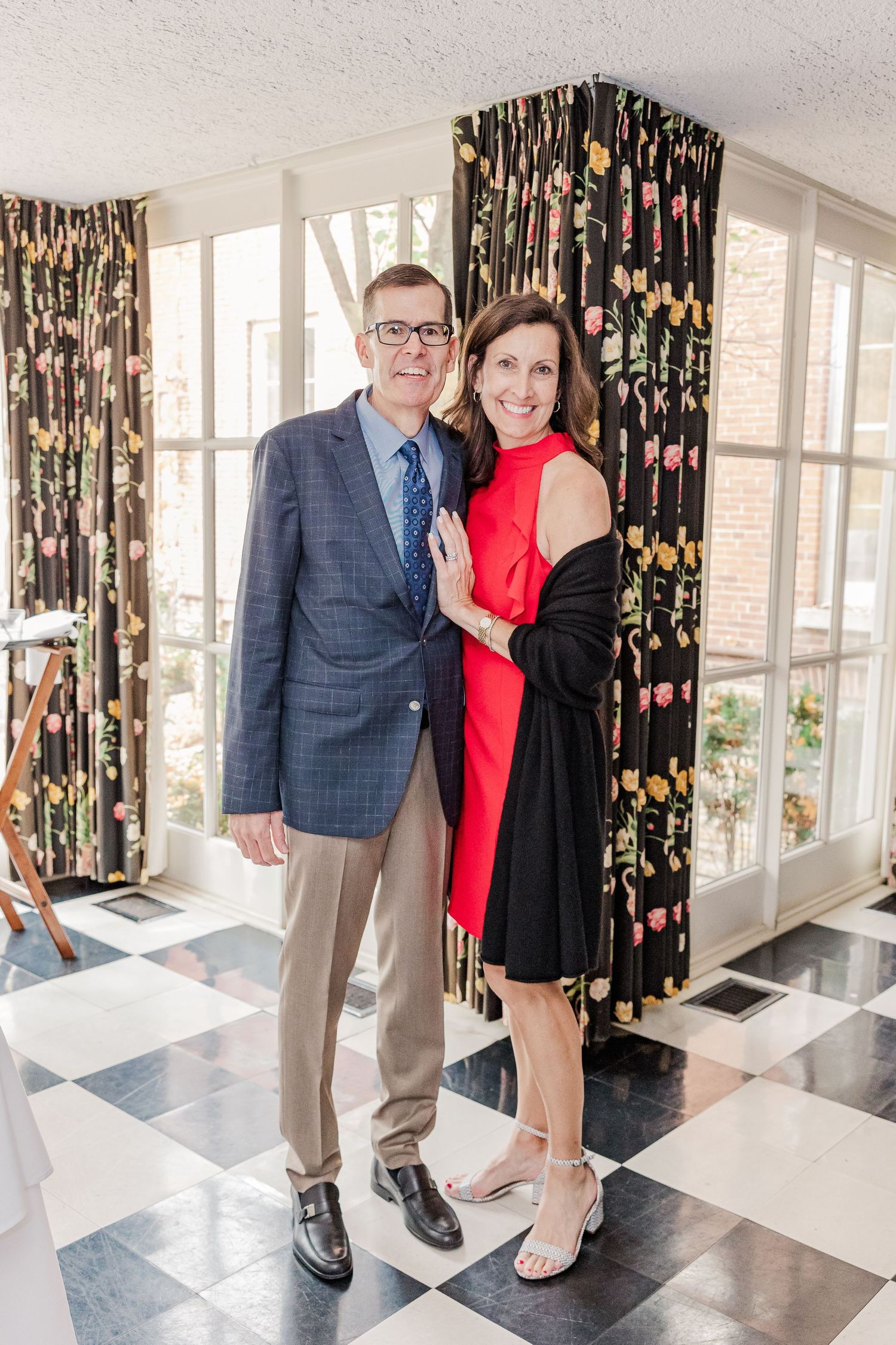A couple poses elegantly in a room with black floral curtains and a checkered floor.