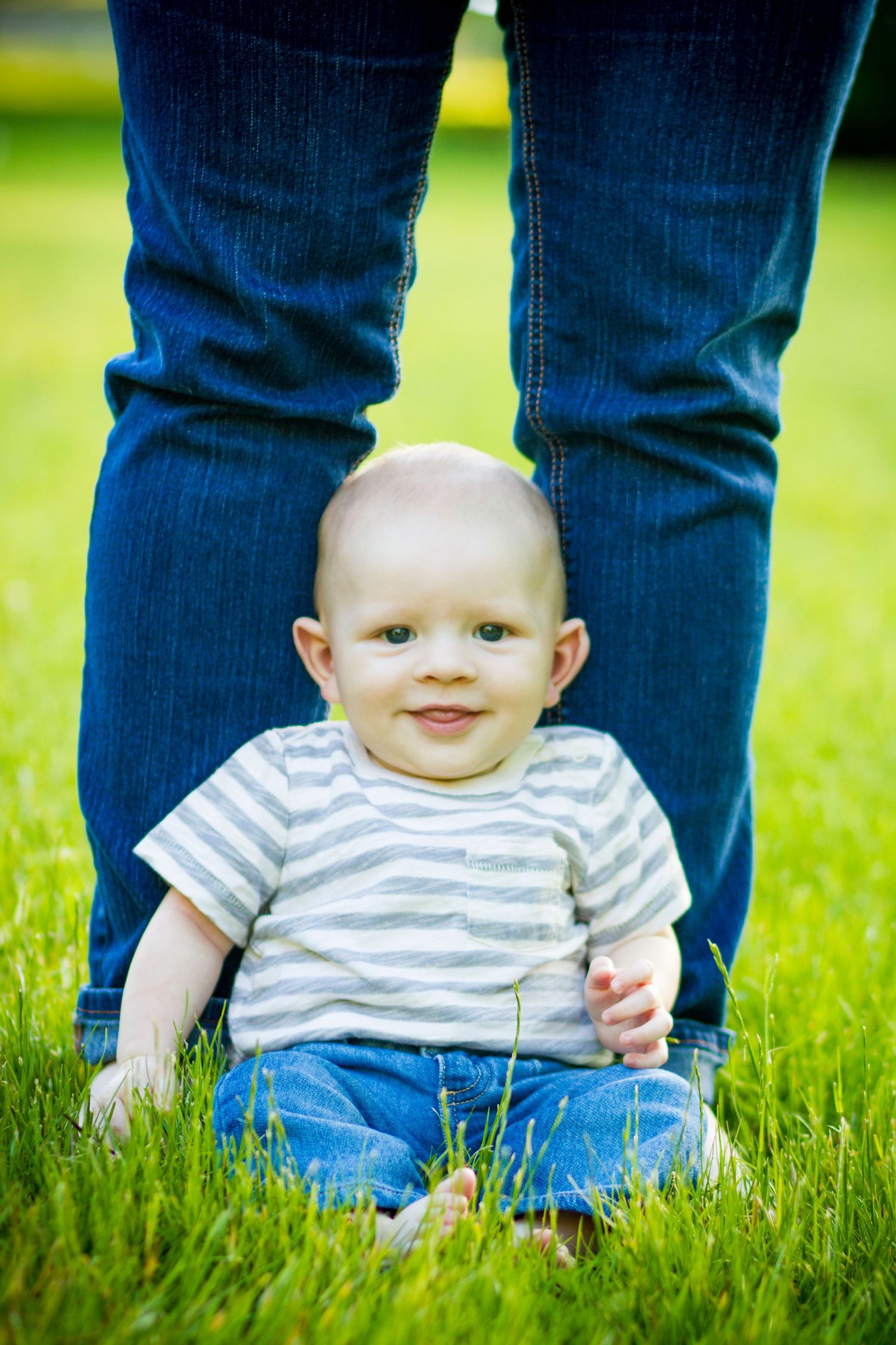 A small child in a striped shirt sits in green grass with adult legs visible behind them.
