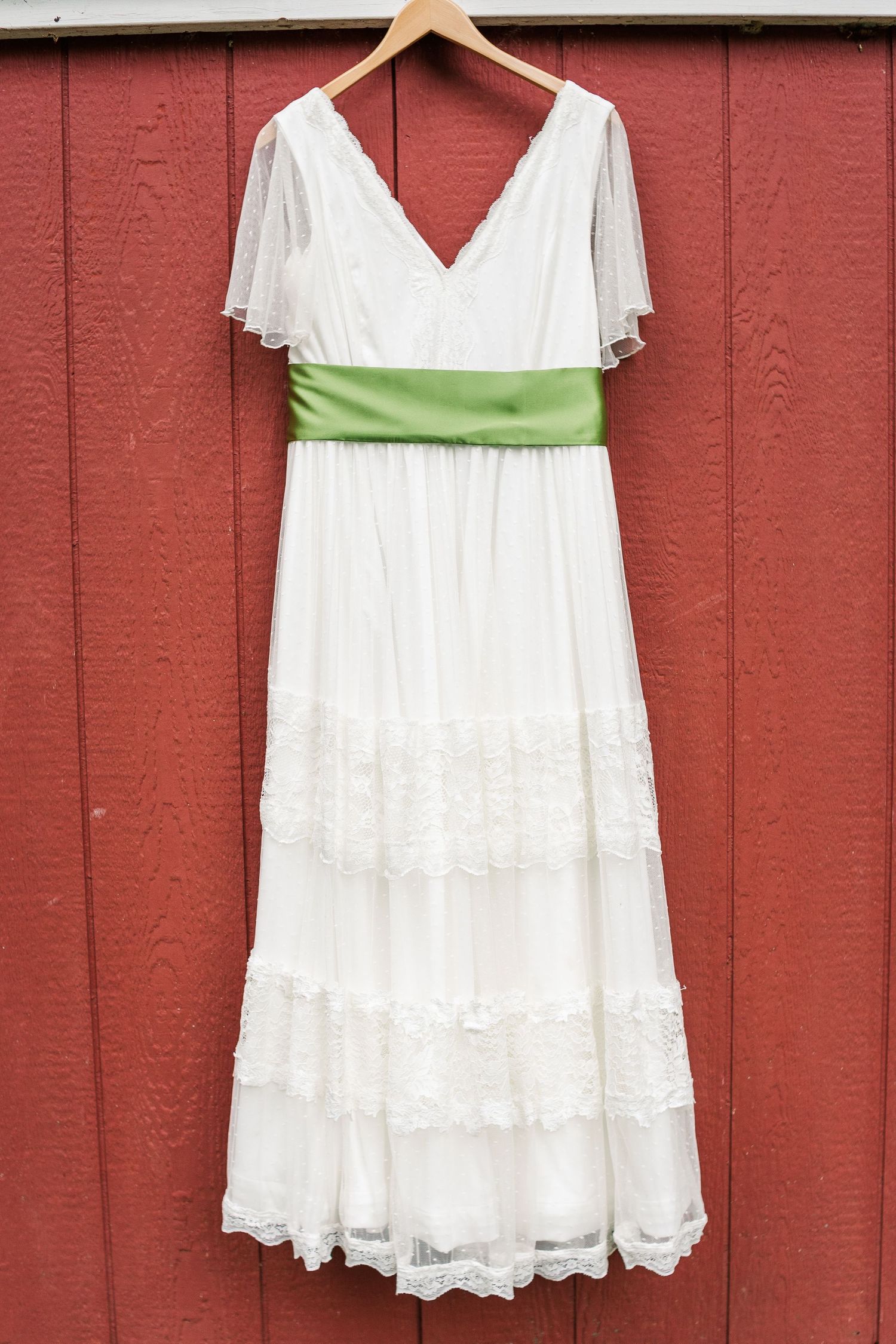 White lace wedding dress with green sash hanging on wooden hanger against red wall.