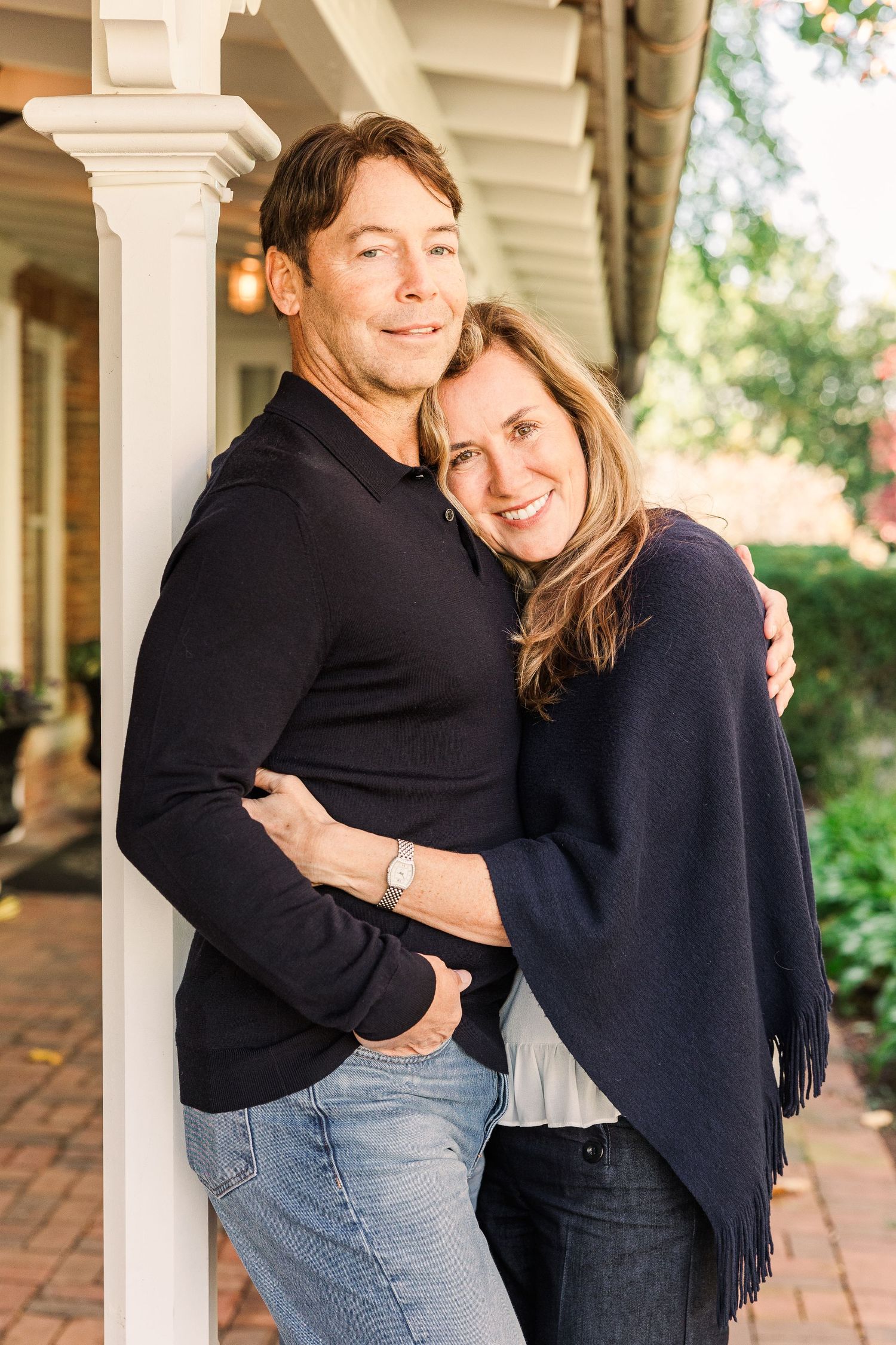 Couple in matching navy sweaters embracing by white porch column on sunny day.