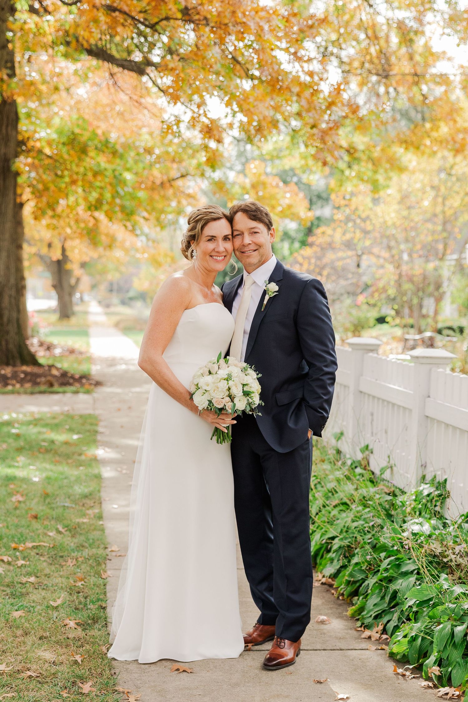 An autumn wedding portrait on a garden path with golden leaves and white fence.