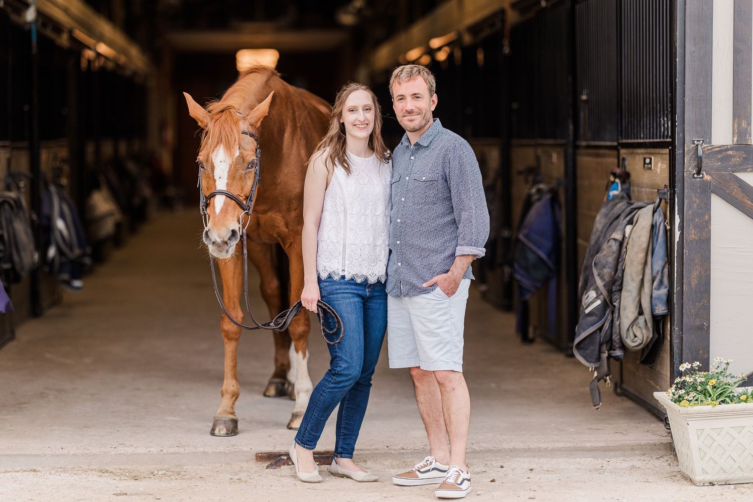 A couple poses with their chestnut horse in a rustic barn setting.