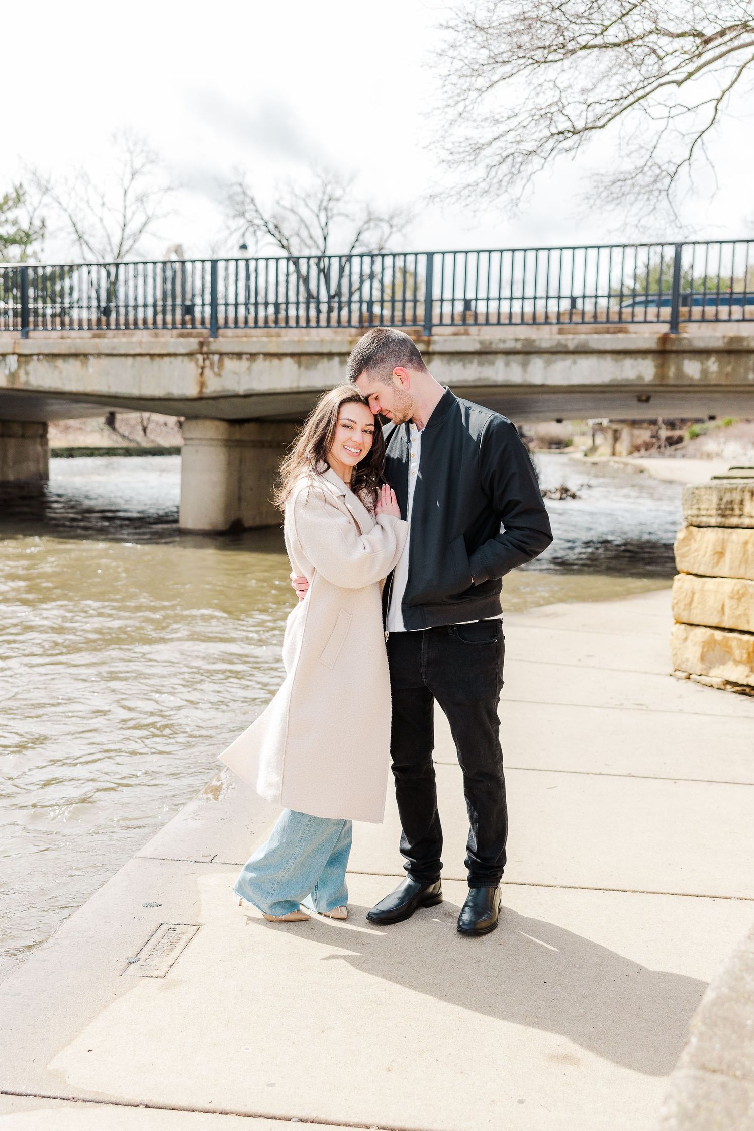 A couple sharing a tender moment by a riverside with a bridge in the background.