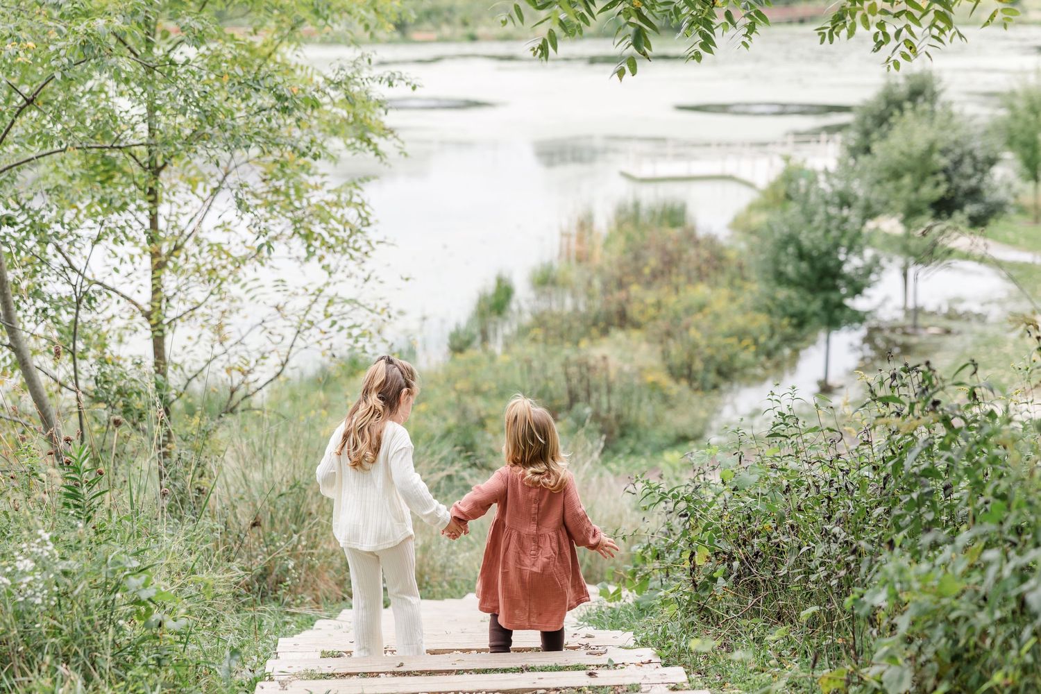 Two young girls holding hands on a wooden path overlooking a serene lake surrounded by lush greenery.