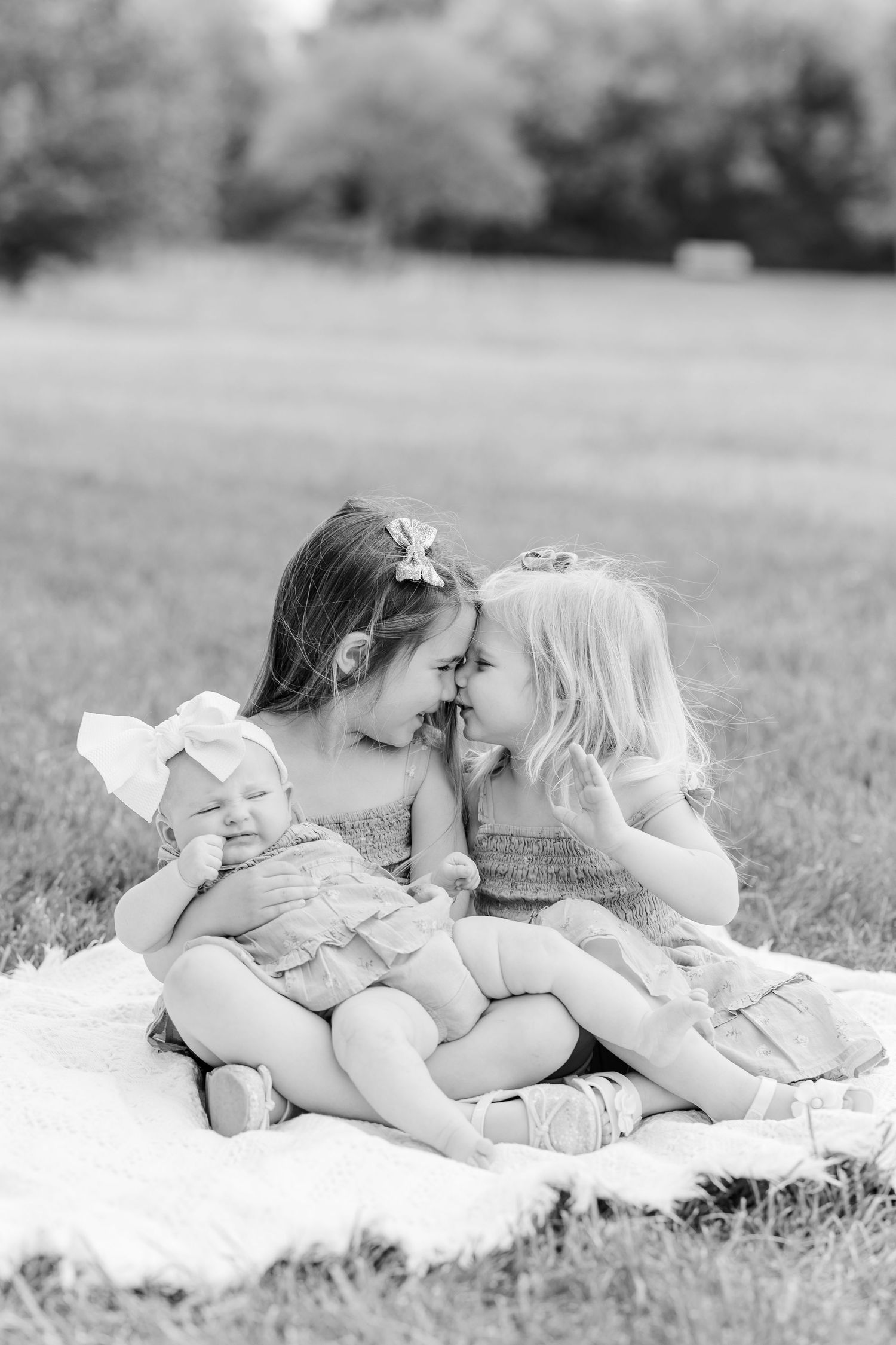 Black and white photo of two young girls sharing a sweet nose-to-nose moment while holding a newborn baby on a blanket outdoors.