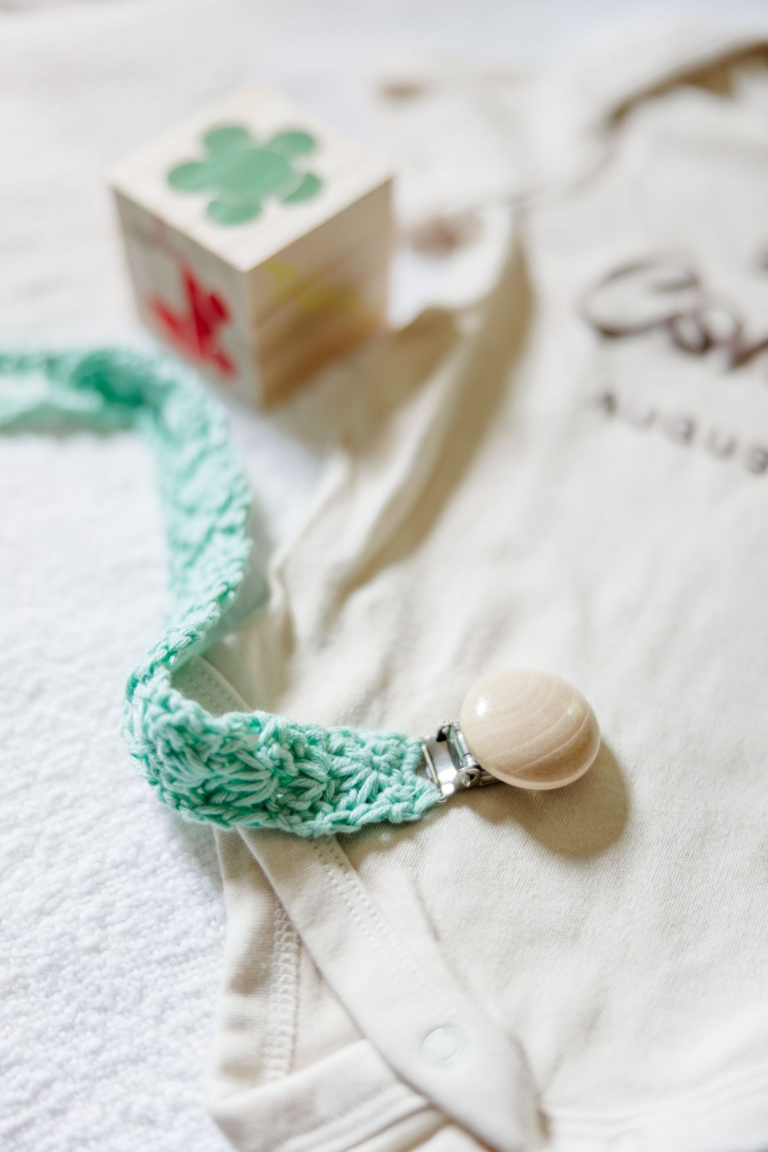 Mint green crocheted pacifier clip with wooden clasp displayed beside a wooden block on a white baby onesie.