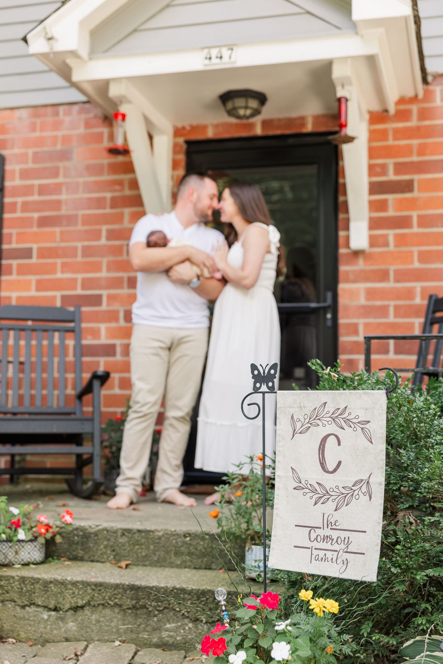 Couple with newborn baby on brick home porch steps beside a Conway family monogram garden flag.