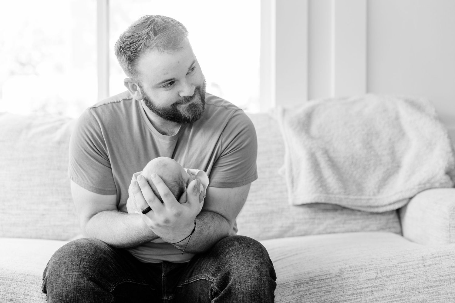 Bearded father holding newborn baby while sitting on couch in black and white photo.