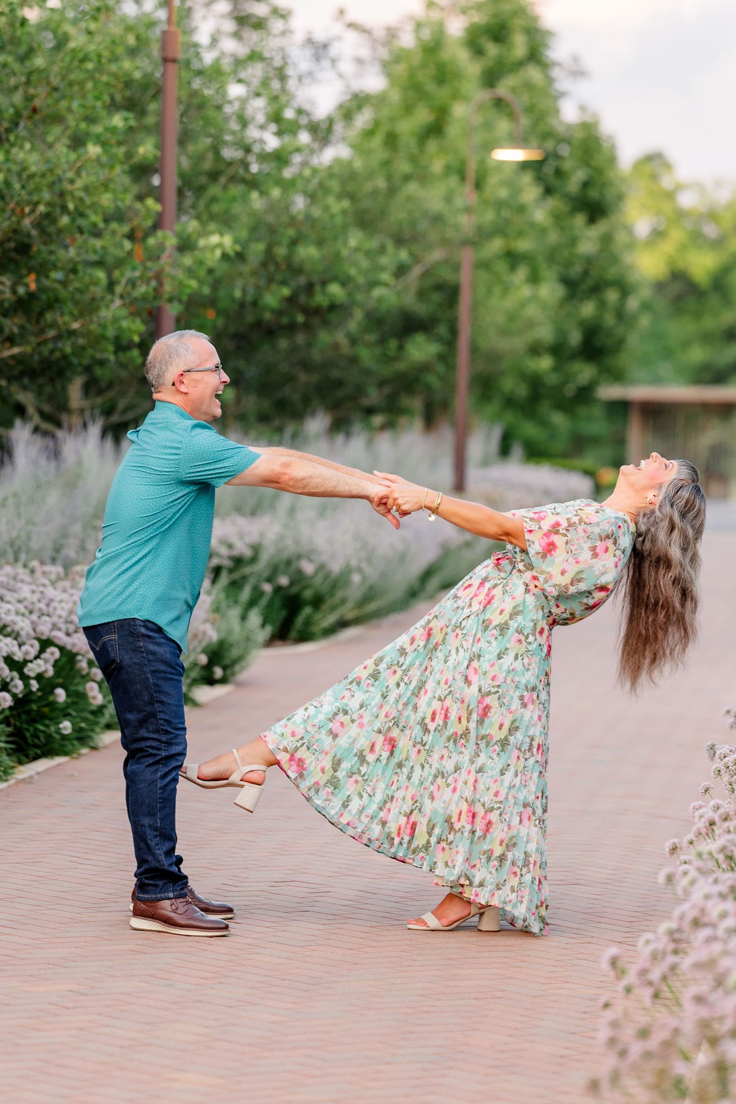 A couple dances joyfully on a garden path, man in teal shirt spinning woman in floral maxi dress outdoors.