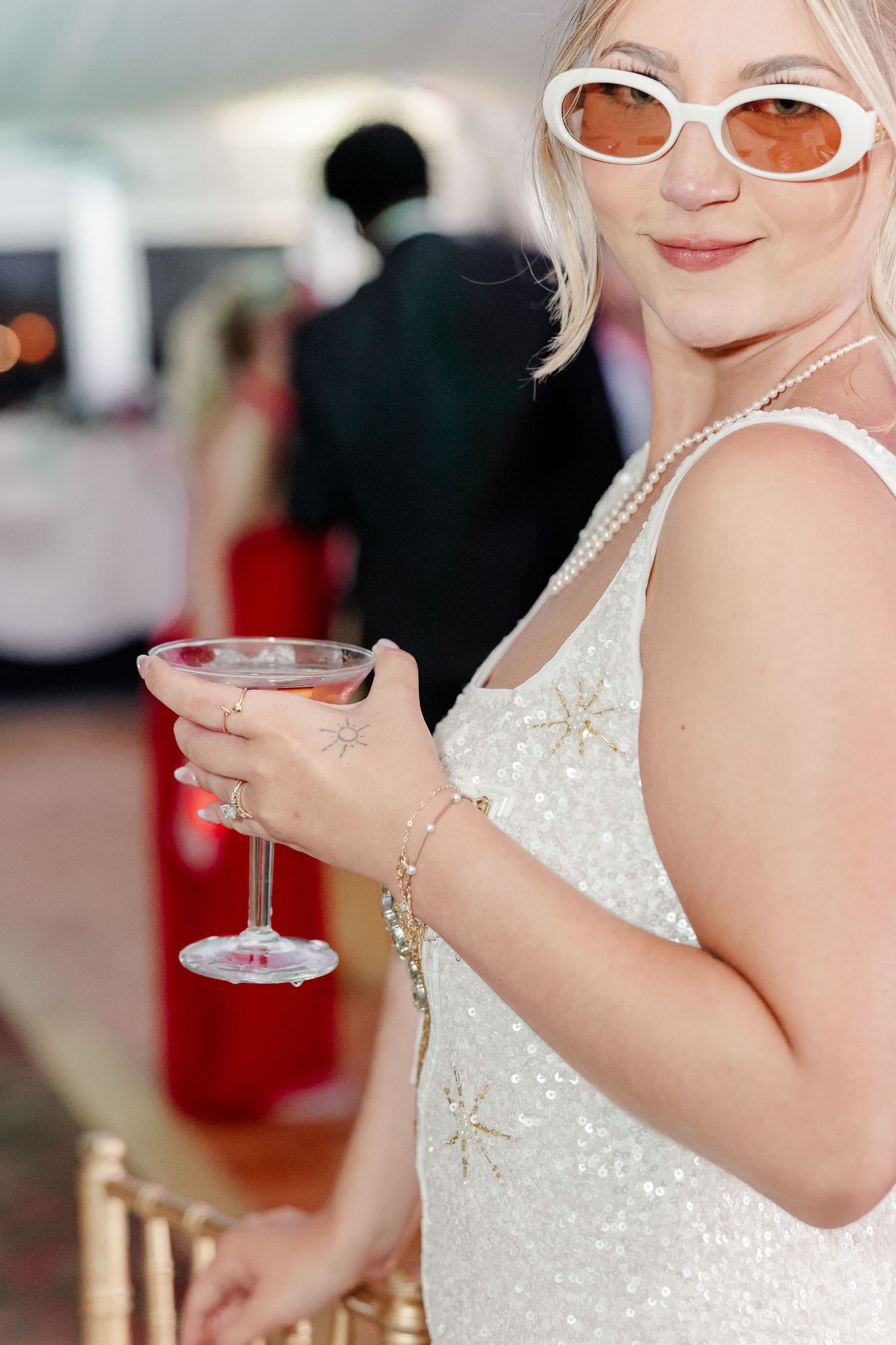 Stylish blonde woman in a sequined white dress and retro sunglasses holds a cocktail at a wedding reception.
