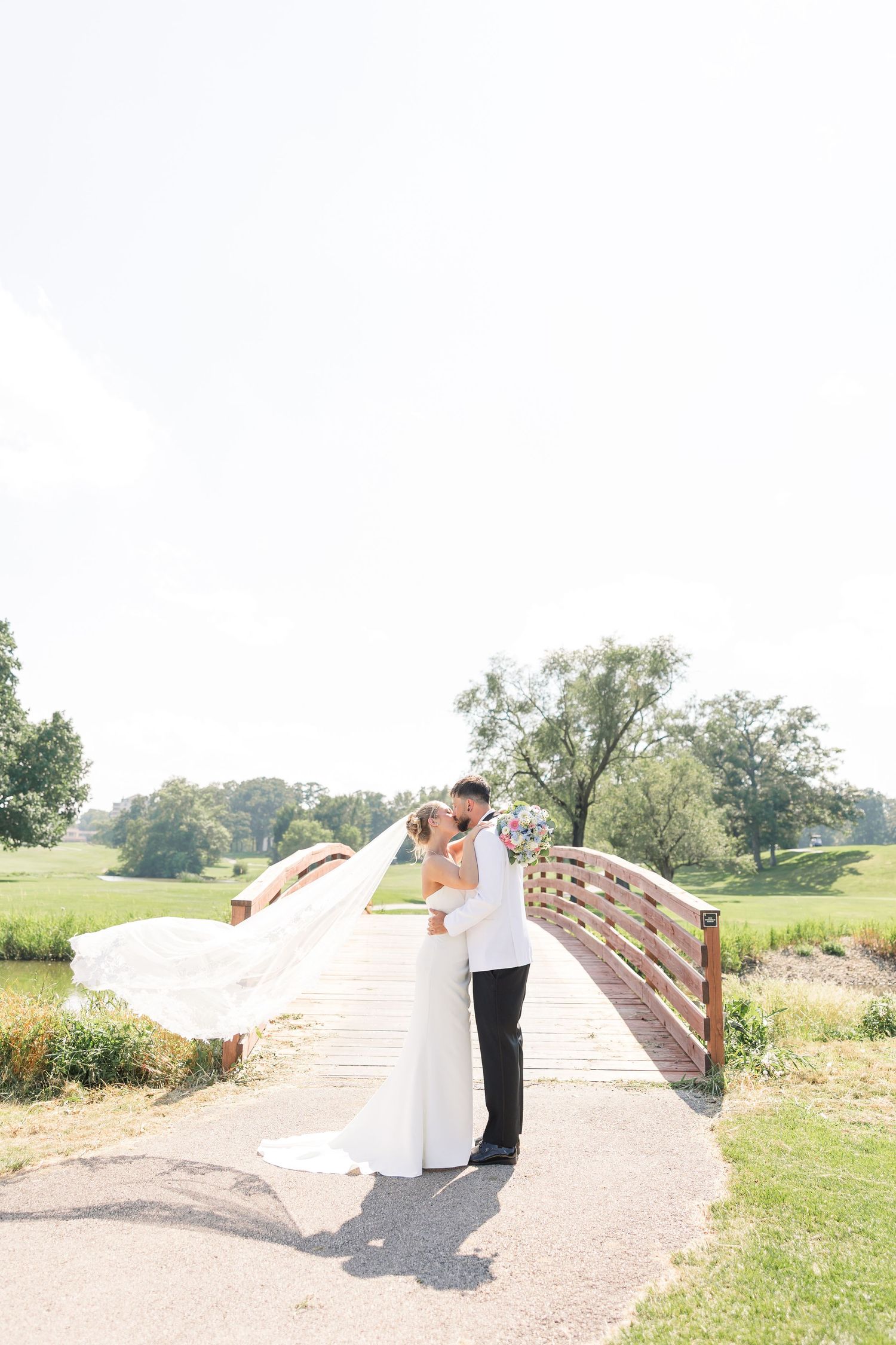 Bride and groom share romantic kiss on wooden bridge with veil flowing in wind at outdoor venue.