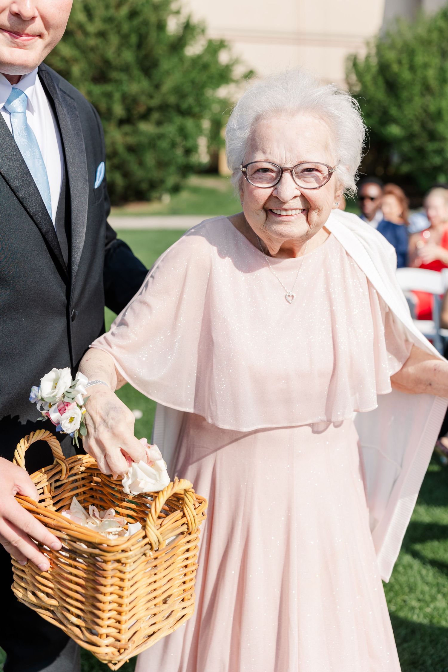 Elderly flower girl in pink dress smiles while holding wicker basket during outdoor wedding ceremony.