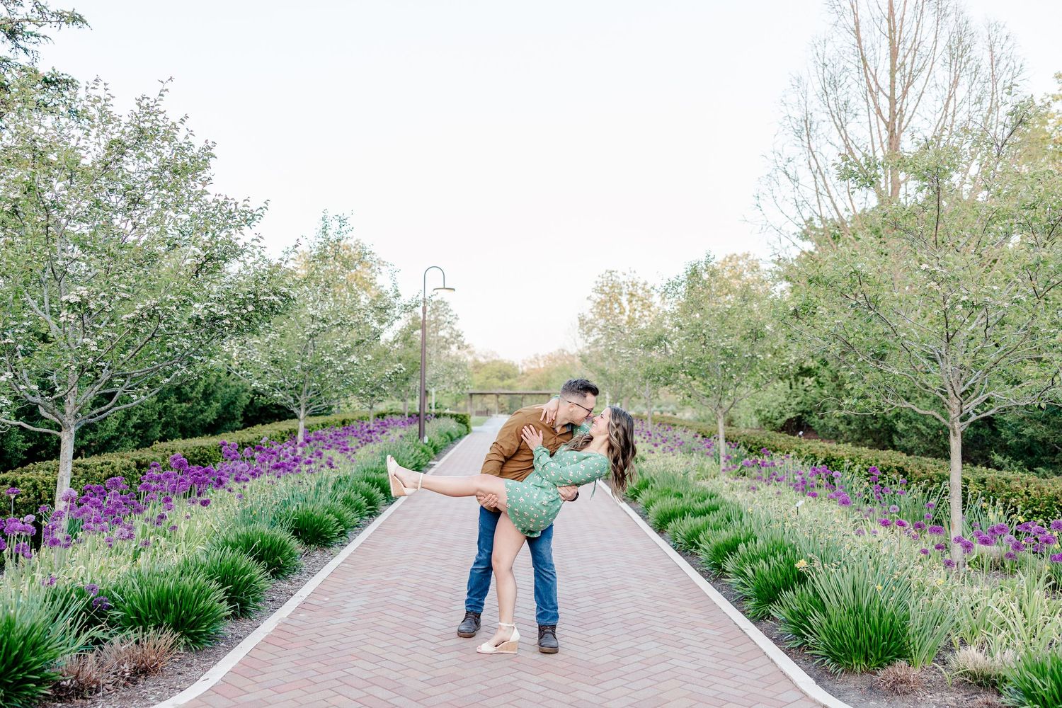 Couple sharing a romantic dip kiss on a brick garden path lined with purple flowers and trees.