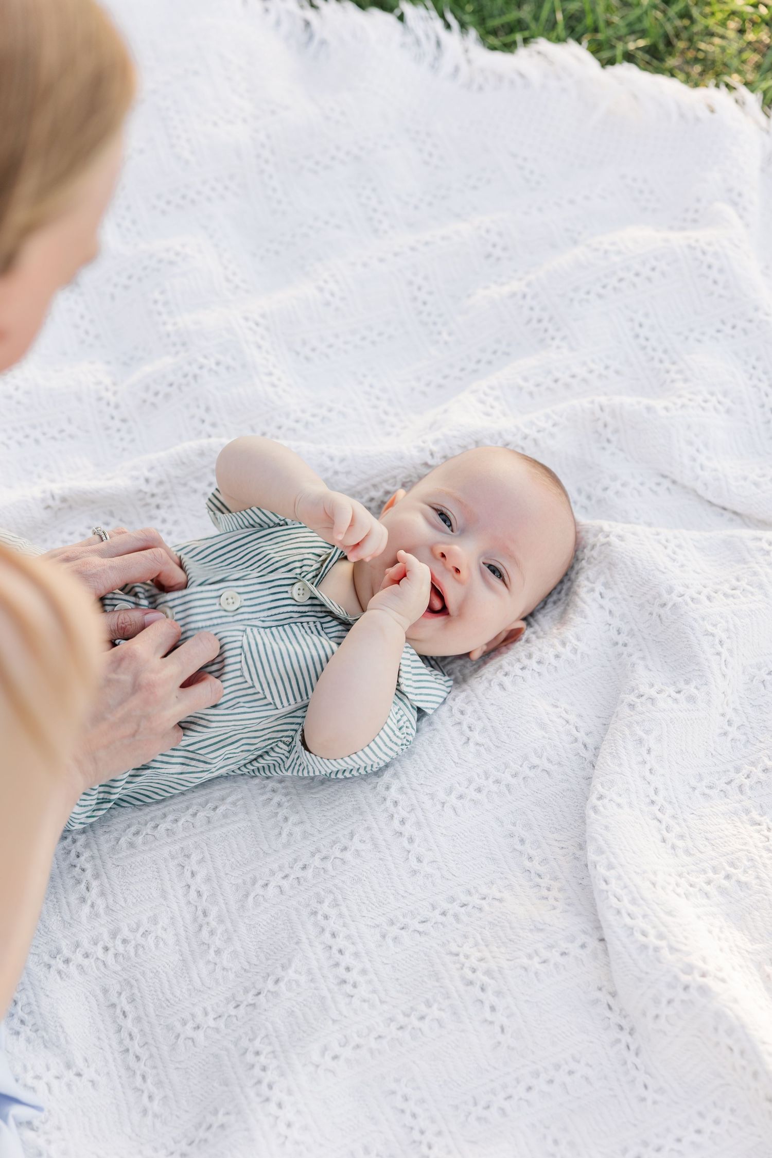 A happy baby in a striped onesie laughs while lying on a white knit blanket outdoors in the sun.