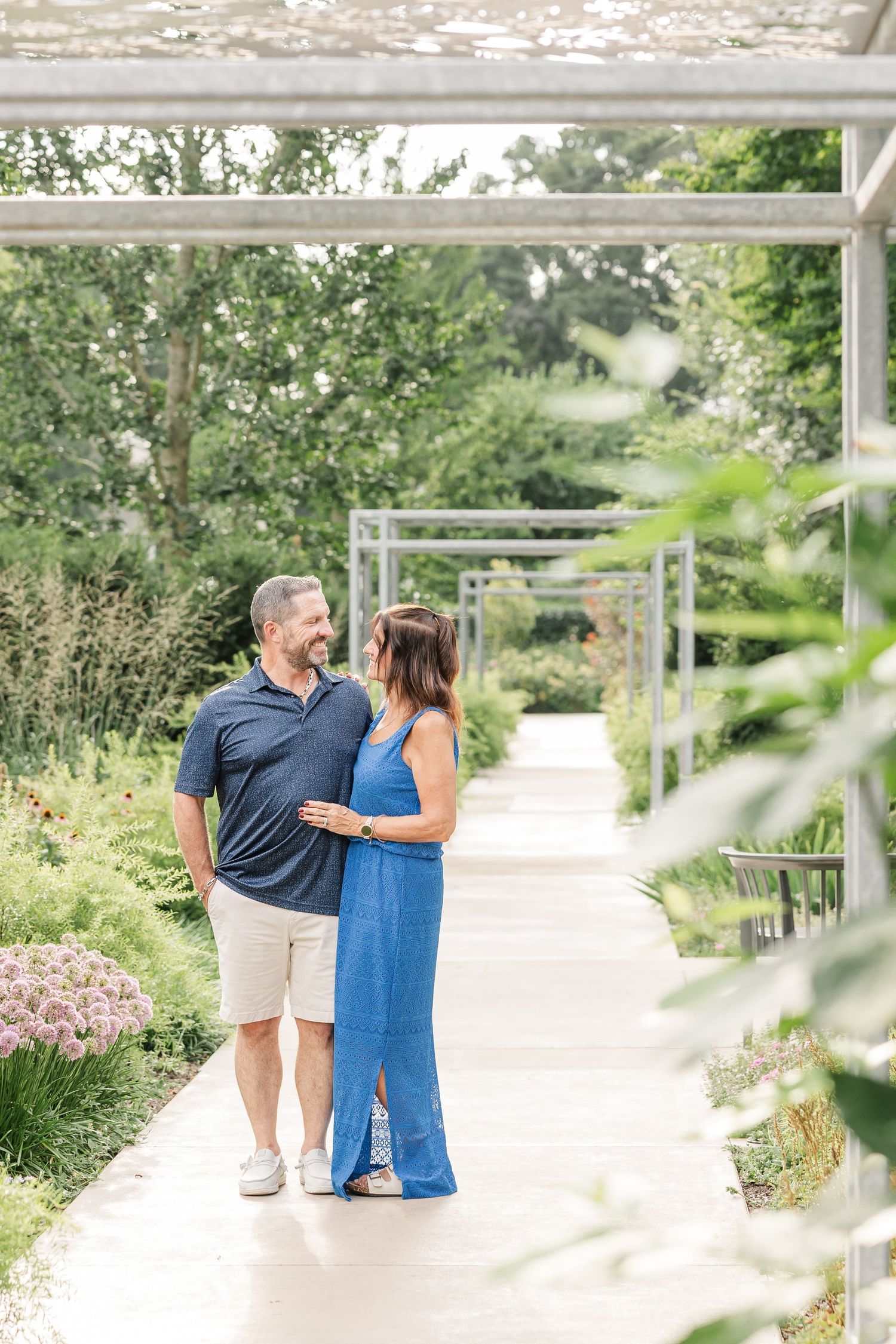 Couple embracing and smiling at each other on a garden pathway surrounded by lush greenery and flowers.