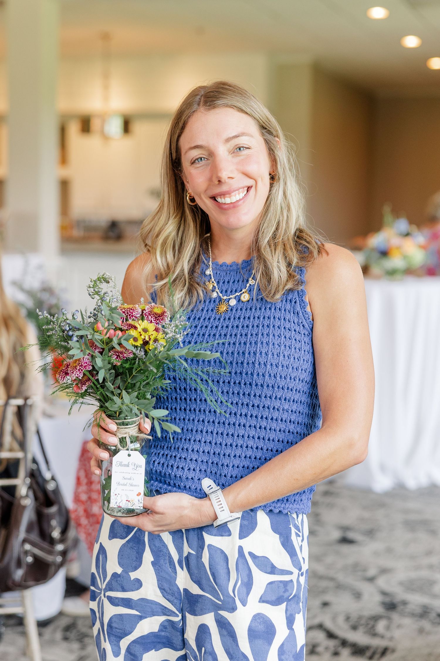 Smiling blonde woman in blue sleeveless top holding a completed wildflower bouquet in a mason jar at a bridal shower.