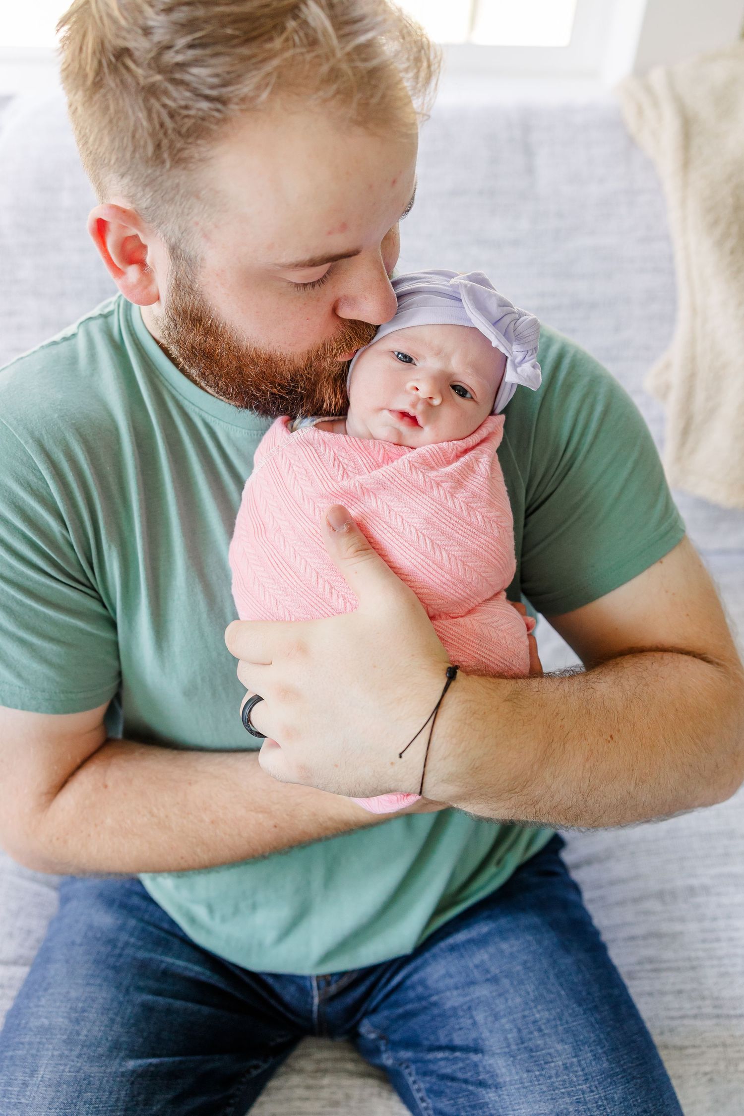 Bearded father in green shirt kissing newborn baby wrapped in pink swaddle blanket with purple hat.
