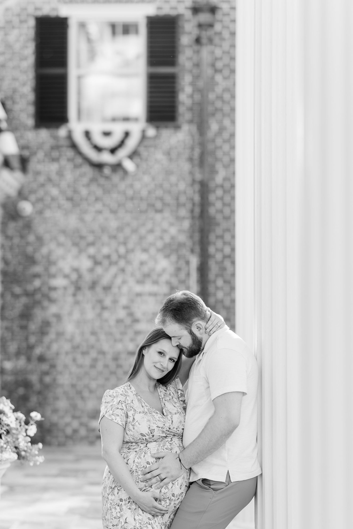 Black and white maternity portrait of parents-to-be cuddling near the brick buildings at Cantigny Park