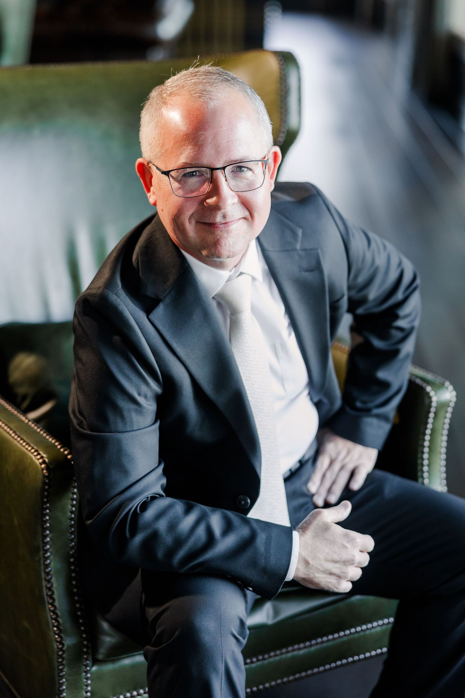 Close-up portrait of a smiling man with glasses wearing a navy suit and light tie, seated in a green studded leather chair.