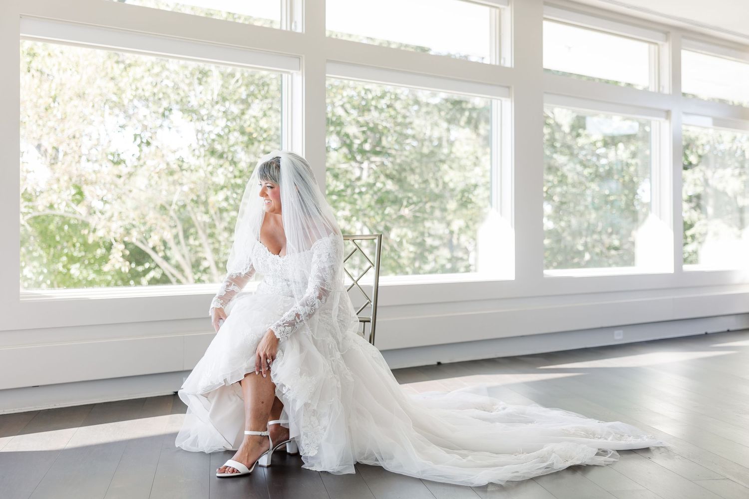 Bride in a lace wedding gown and veil adjusts her white strappy heel while seated near bright windows with leafy views.