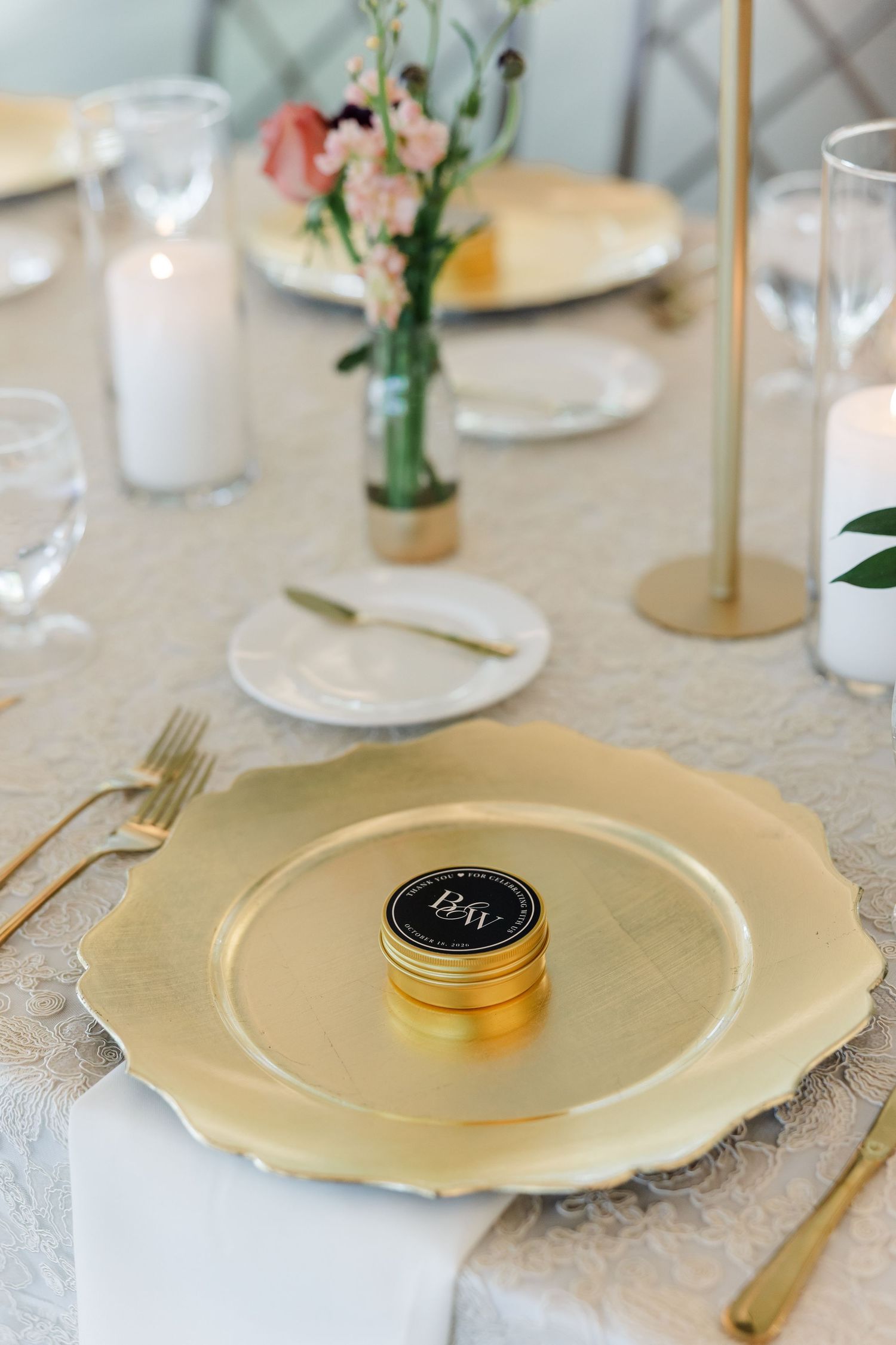 Gold scalloped charger plate with a small black favor tin, gold flatware, white napkin and a pink floral vase at an elegant wedding table.