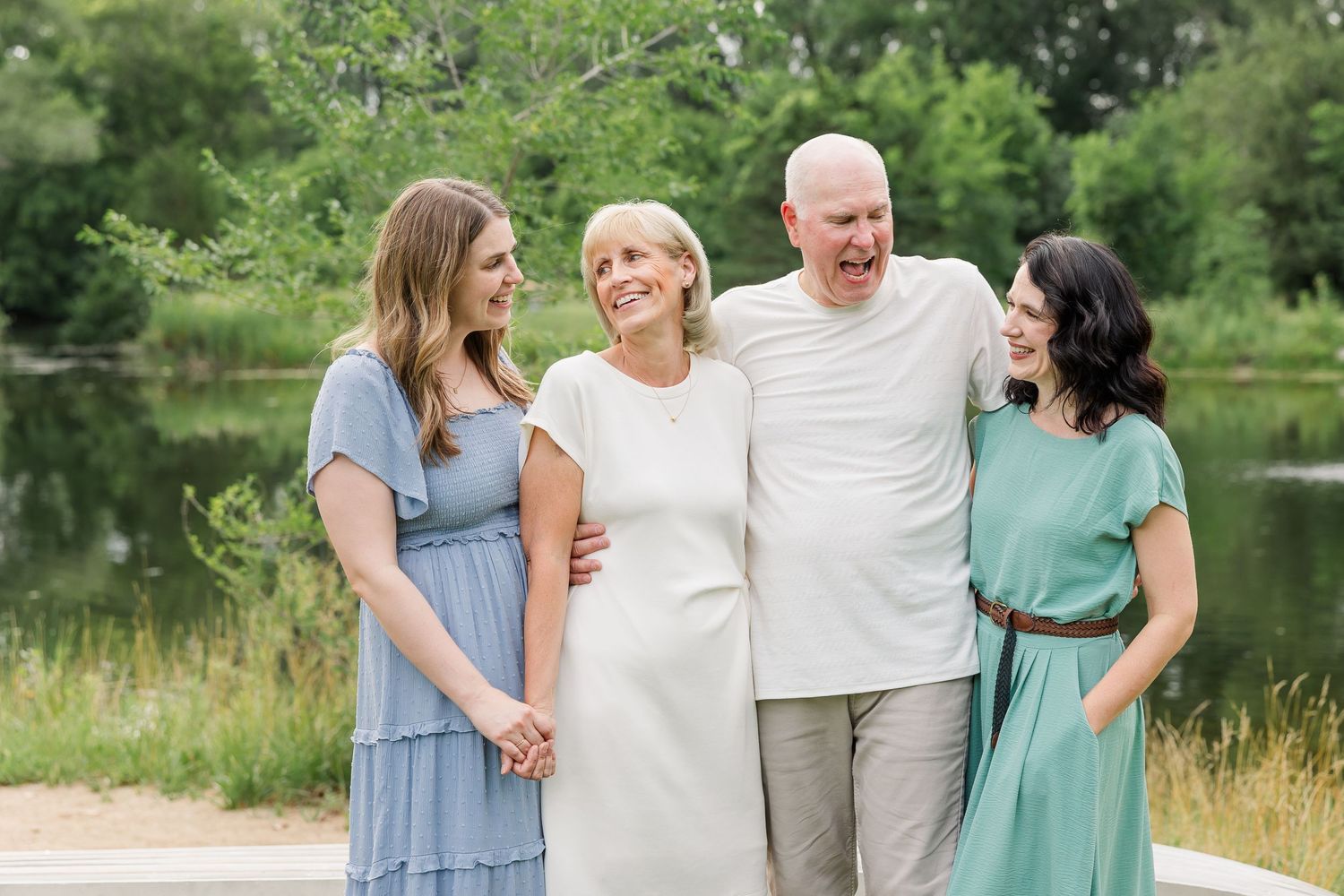 Happy family of four smiling outdoors near a pond, wearing pastel dresses and casual attire in a lush green setting.