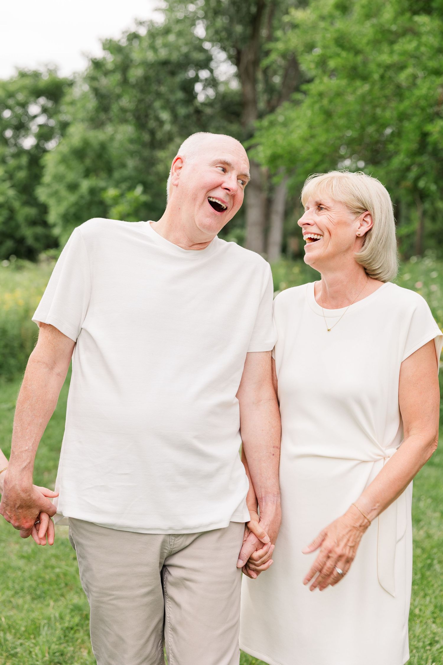 Smiling elderly couple holding hands and laughing together outdoors in a lush green park setting.