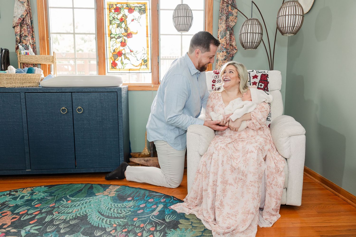 A man kneels beside a woman in a floral dress seated in a white chair, sharing a tender moment in a cozy home.