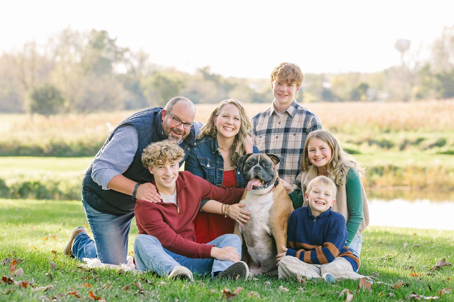 Smiling family of six posing with their boxer dog on a grassy lawn by a pond during a sunny outdoor autumn portrait session.