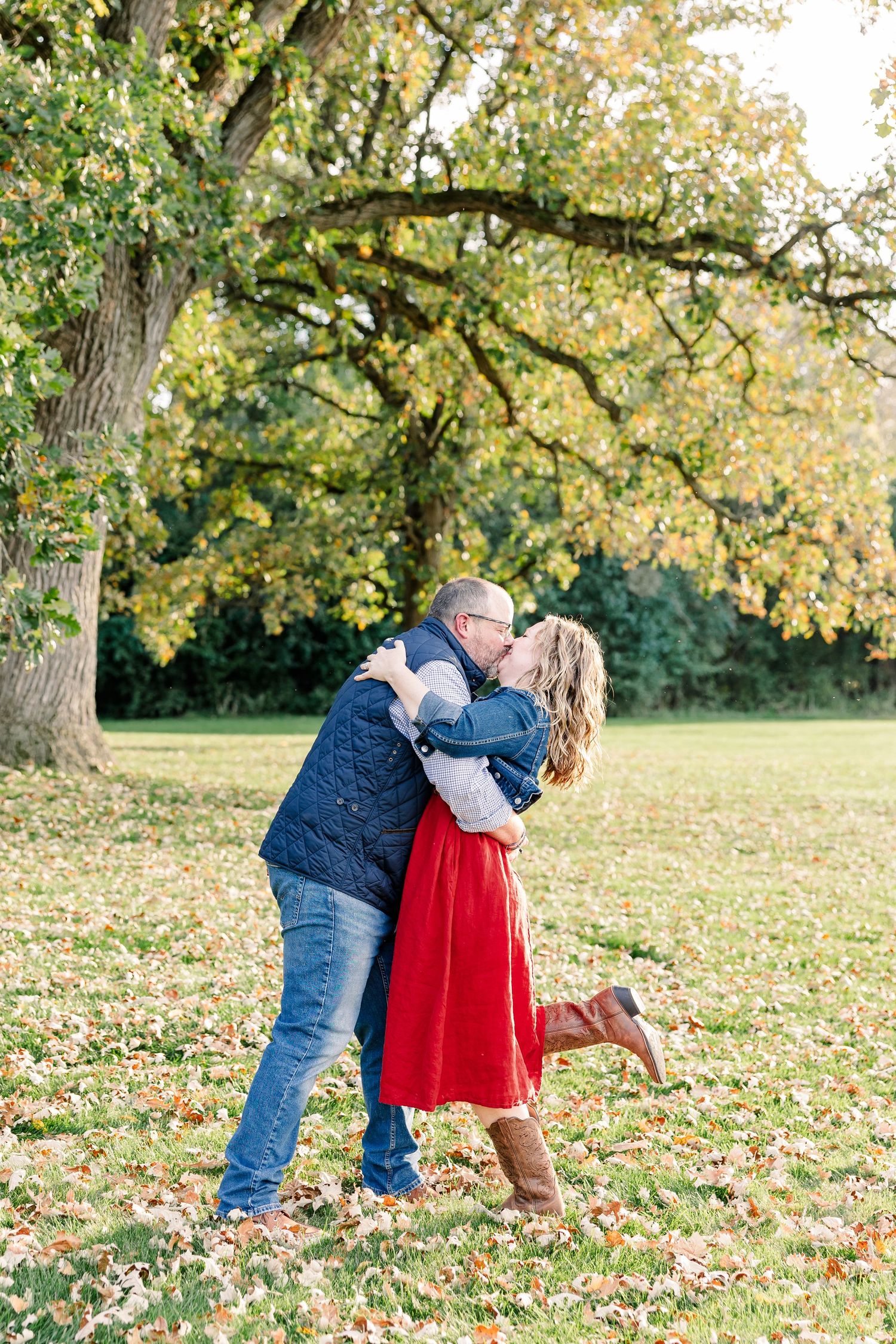 Couple shares a joyful kiss beneath a large autumn tree as the woman in a red skirt and brown boots lifts one foot.