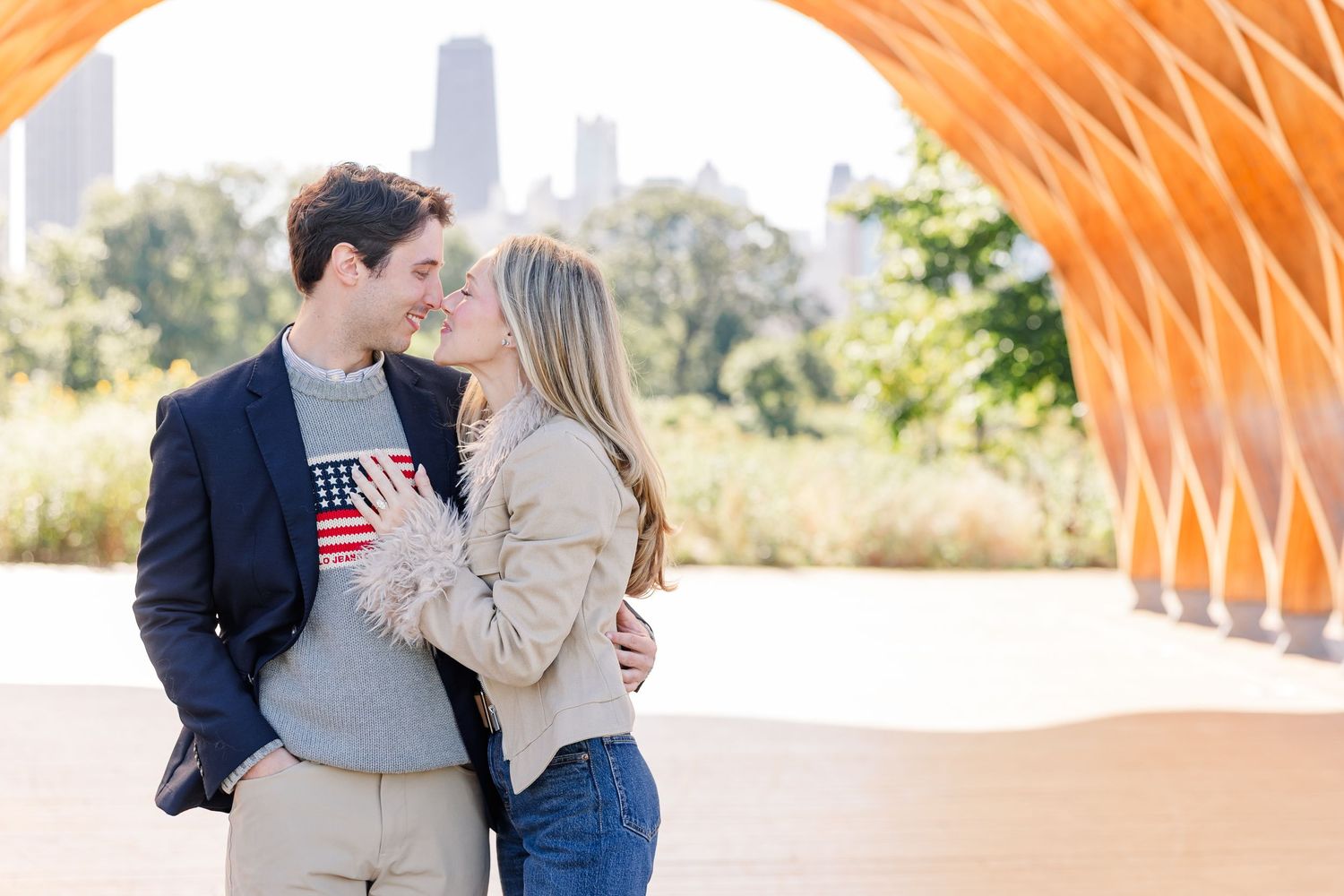 Couple embracing and about to kiss outdoors near an orange architectural structure with city skyline in background.