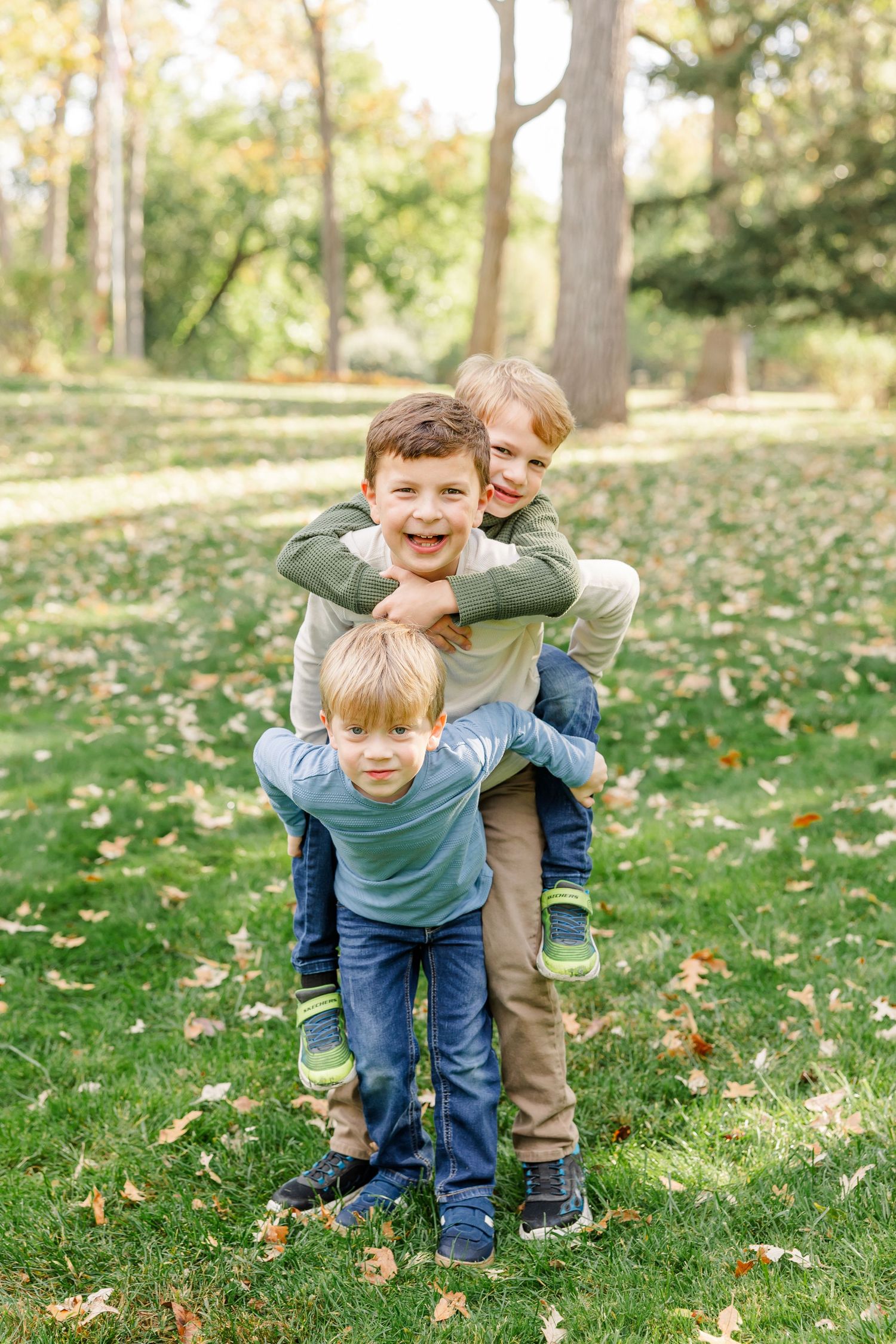 Three brothers stack up playfully on green grass in an autumn park, smiling for a family photo.