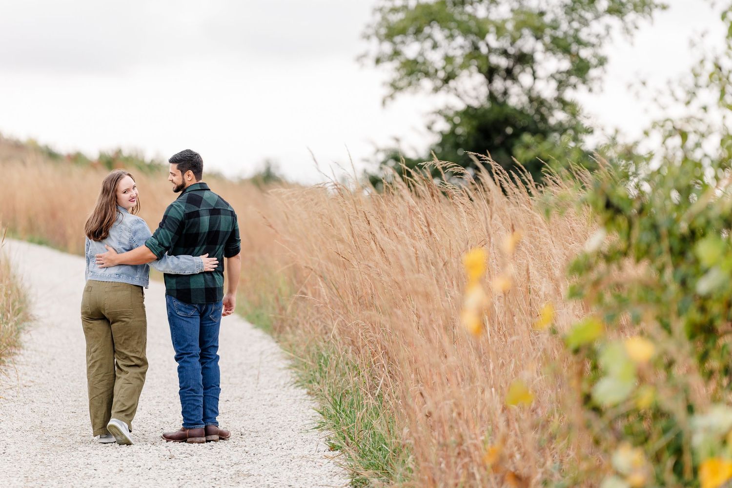Couple in casual clothes walking arm in arm down a gravel path through golden prairie grass with trees in the background.