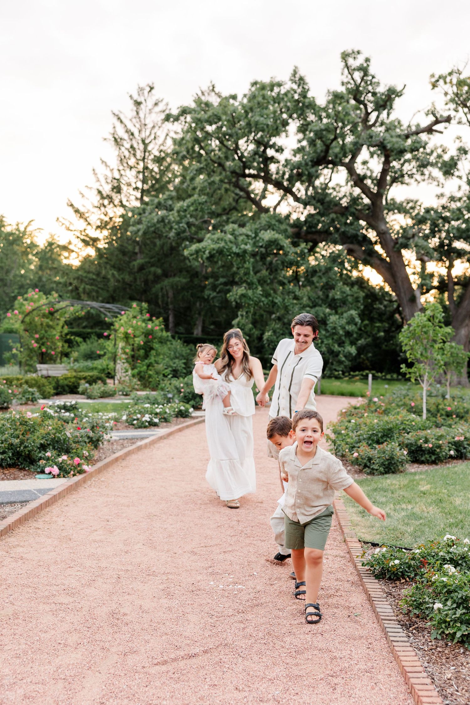 Joyful family walks along garden path at sunset as young boy runs toward camera with excitement.