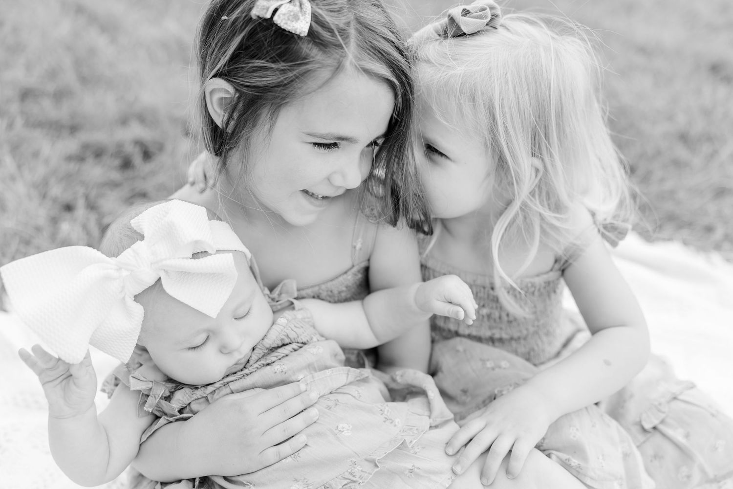 Two young sisters in dresses tenderly holding their newborn baby sibling on a white blanket, black and white photo.