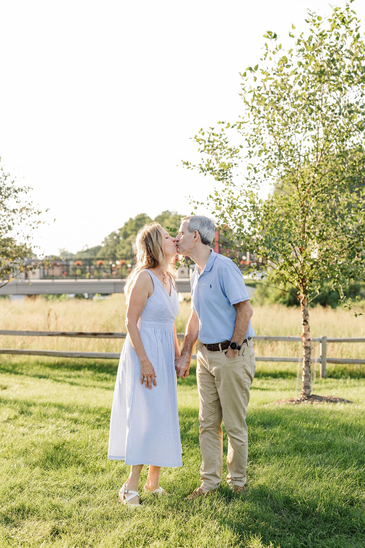 Middle-aged couple kissing while holding hands outdoors in green park near wooden fence.