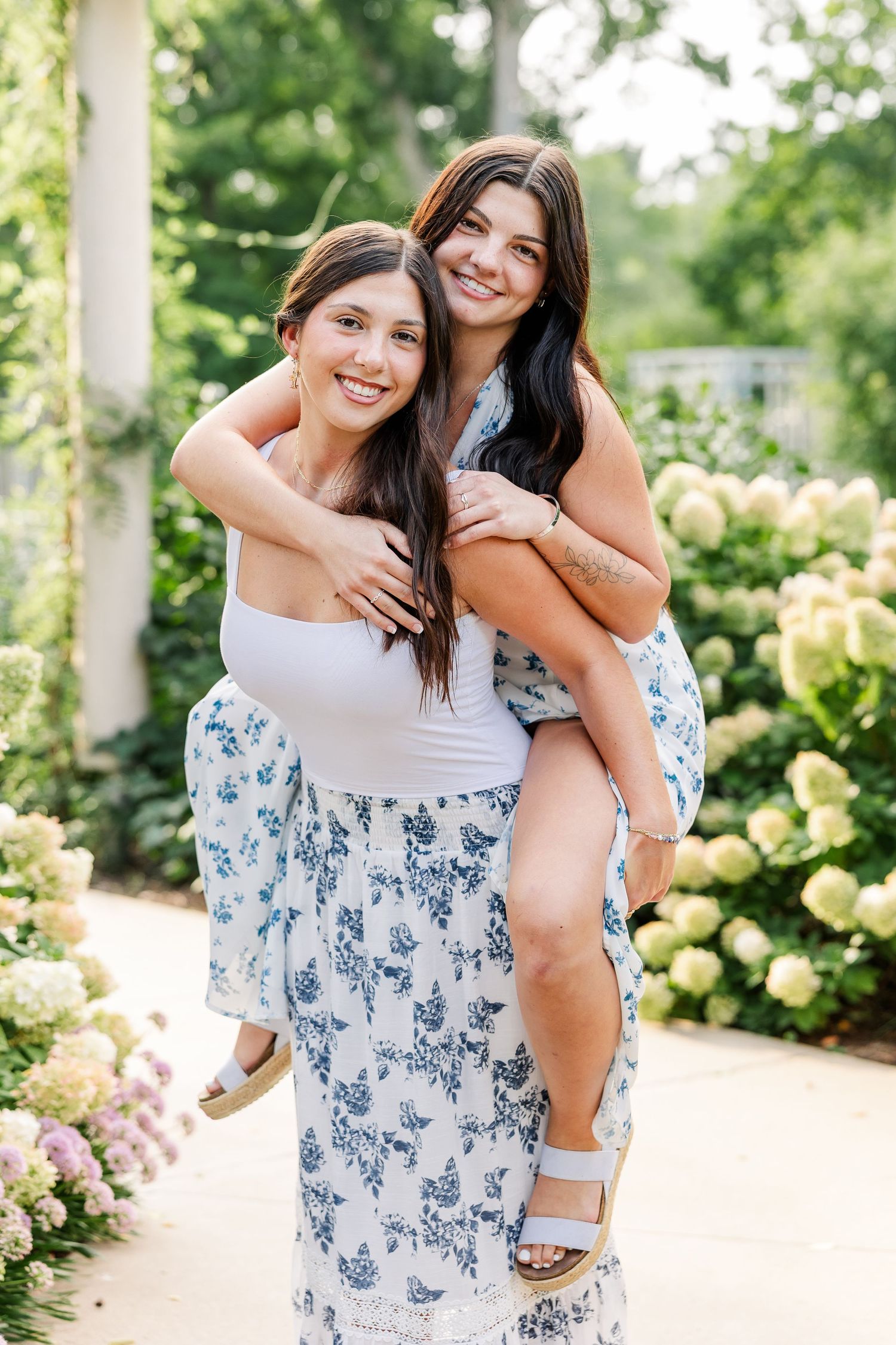 Two brunette sisters smile as one gives the other a piggyback ride in garden.