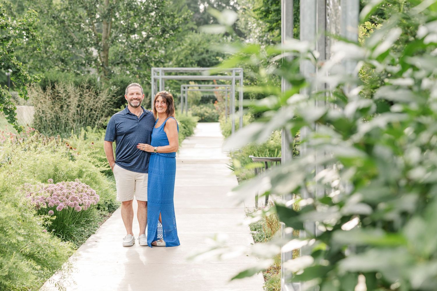 Couple poses on garden path surrounded by lush greenery, woman in blue dress and man in navy shirt, outdoor engagement photo.