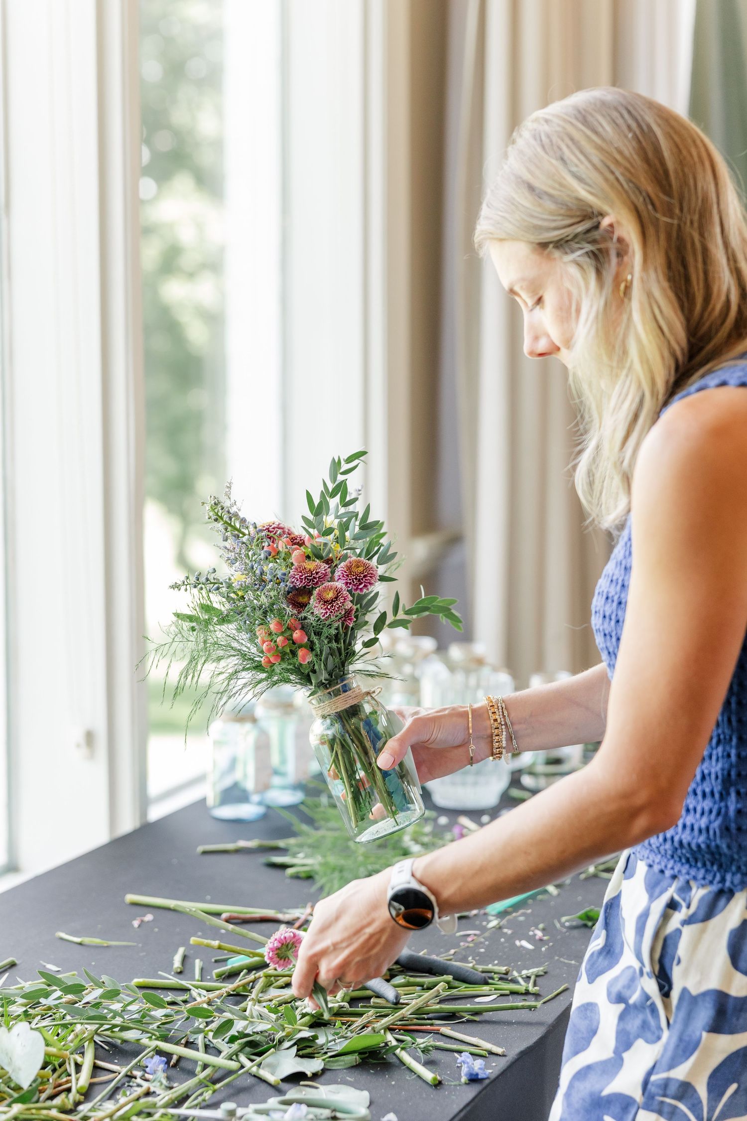 Blonde woman in blue floral dress placing a pink and green wildflower bouquet into a mason jar at a flower arranging table.
