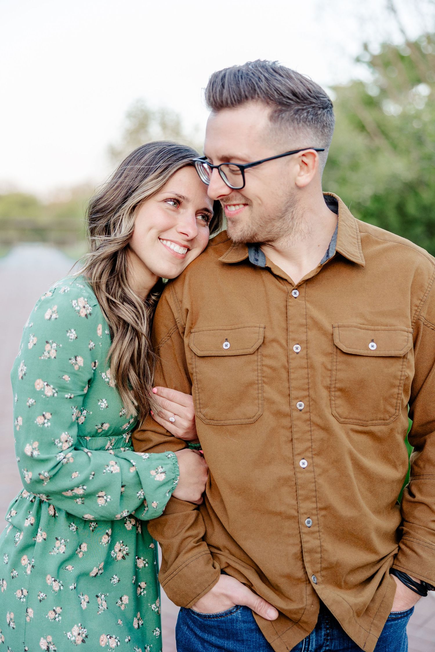 A smiling couple in a romantic outdoor portrait, woman in green floral dress leaning on man in brown shirt wearing glasses.