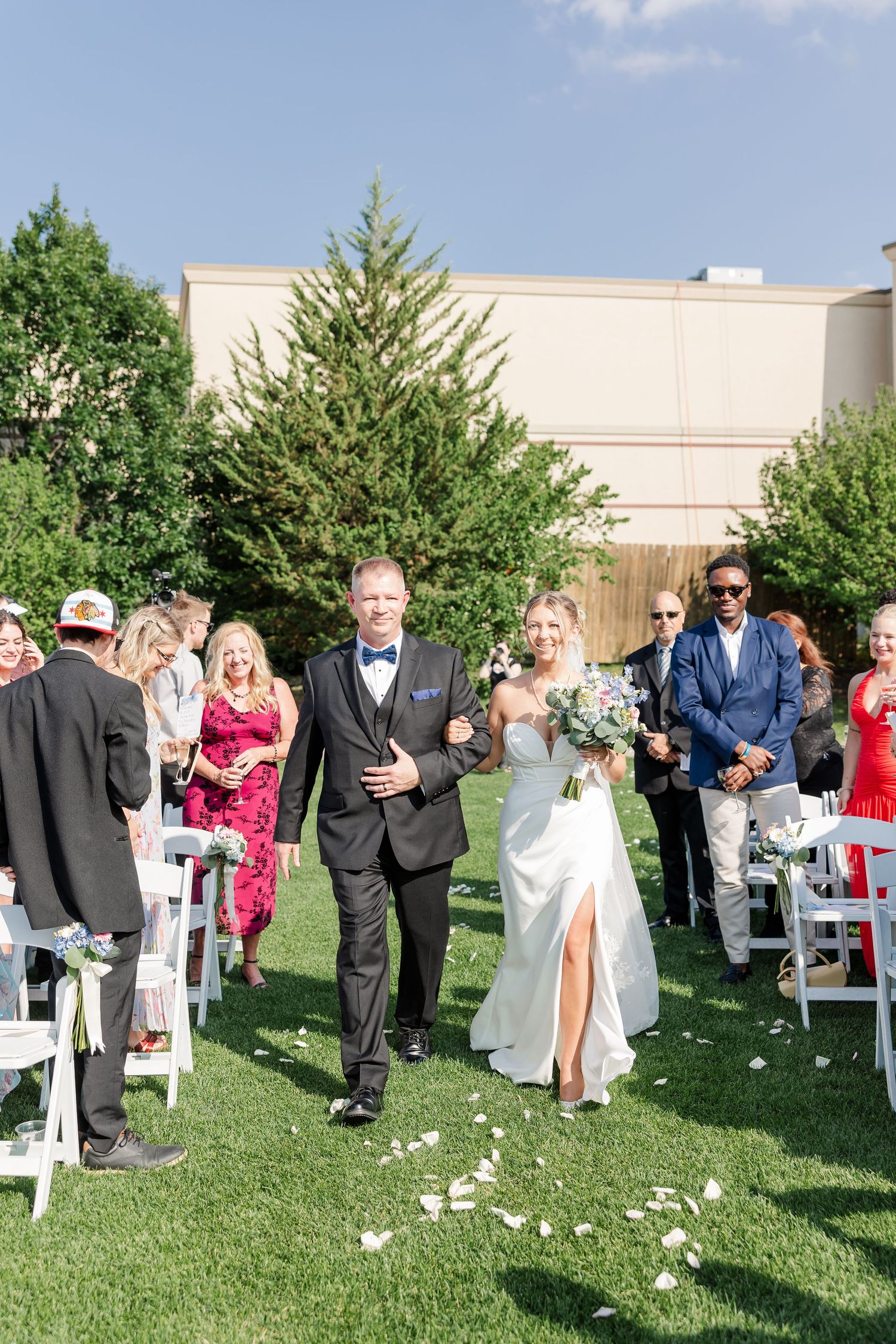 Bride and father walking down outdoor aisle after ceremony, guests celebrating with white floral petals.