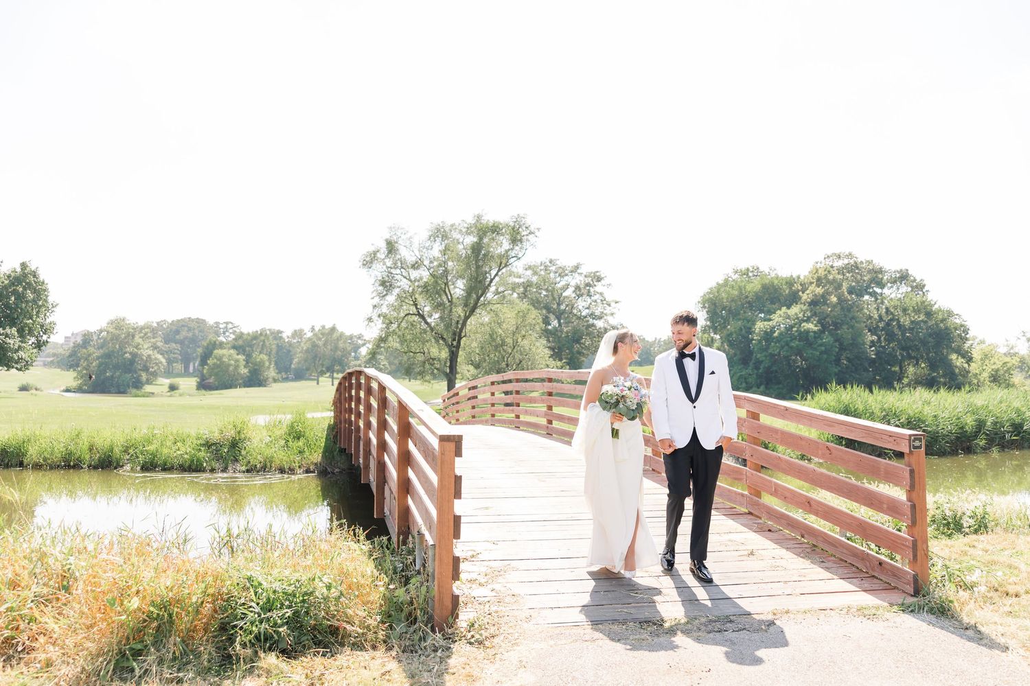 Bride and groom walking hand in hand on a wooden bridge surrounded by green fields.