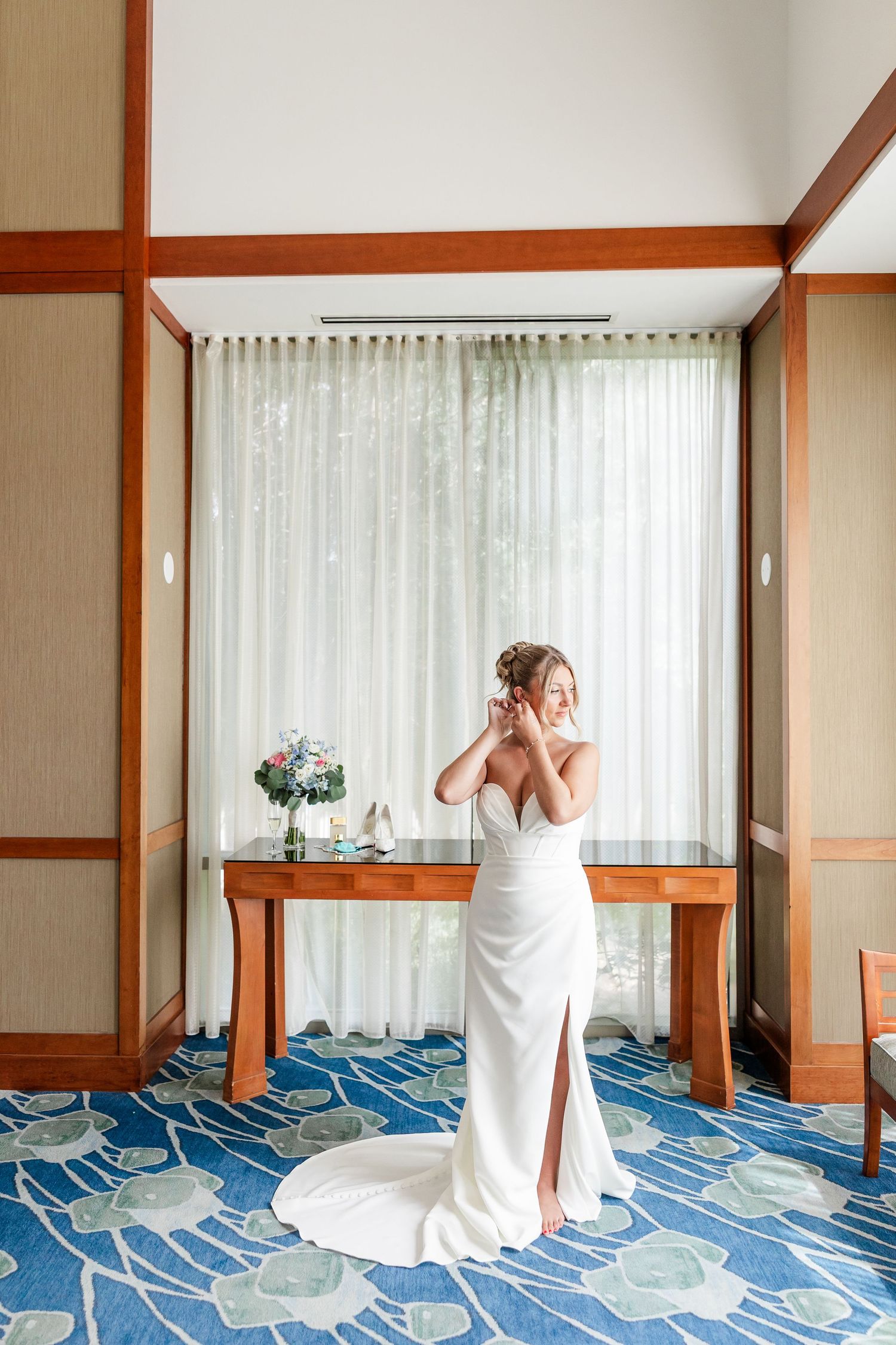 Bride in a white fitted wedding gown adjusting her earring near a window in an elegant hotel room.