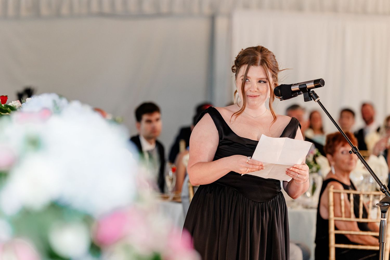 A bridesmaid in a black gown delivers a heartfelt speech at a wedding reception into a microphone.