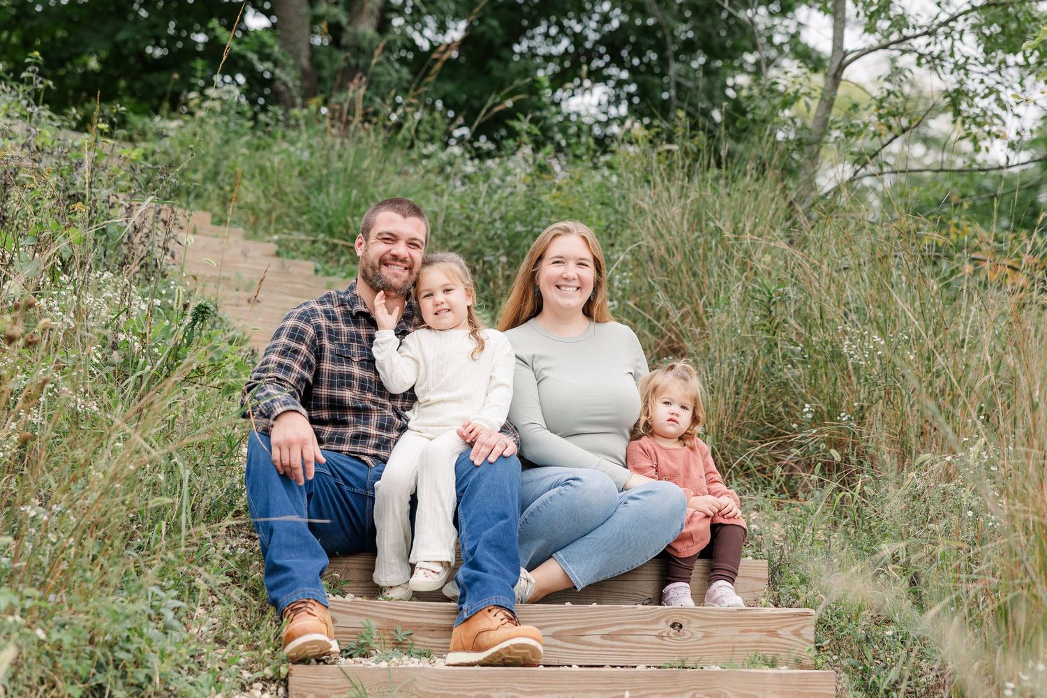 Family of four smiling on wooden steps surrounded by lush greenery during an outdoor fall photo session.