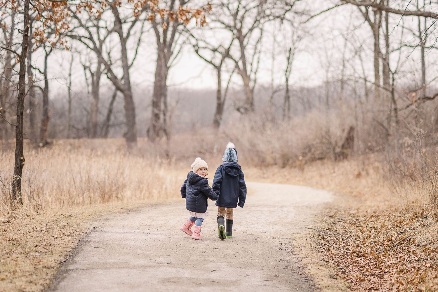 Two small children walk together down a winding path through autumn woods wearing warm winter coats.