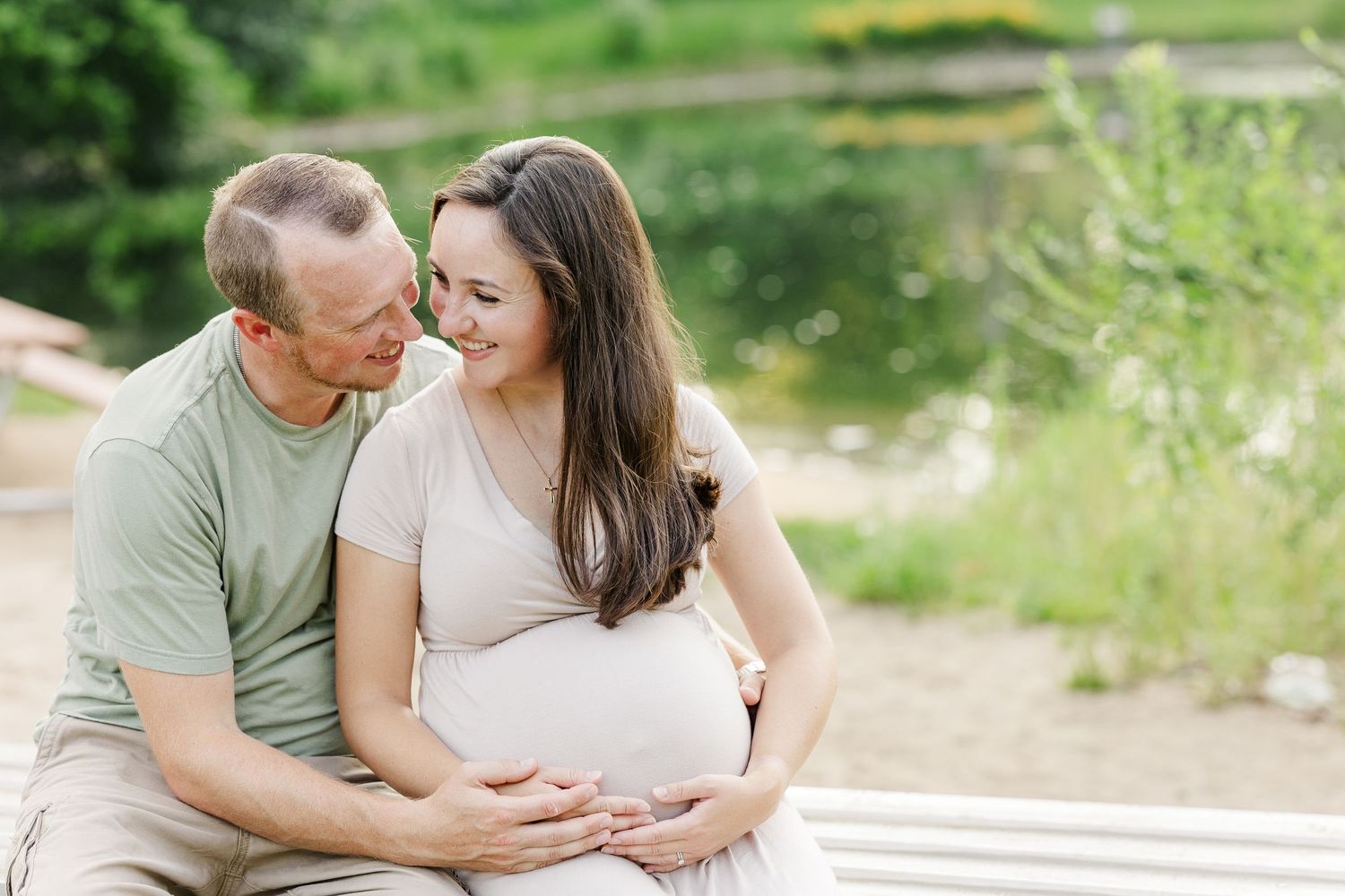 Expecting couple shares intimate moment on bench near pond surrounded by summer greenery.